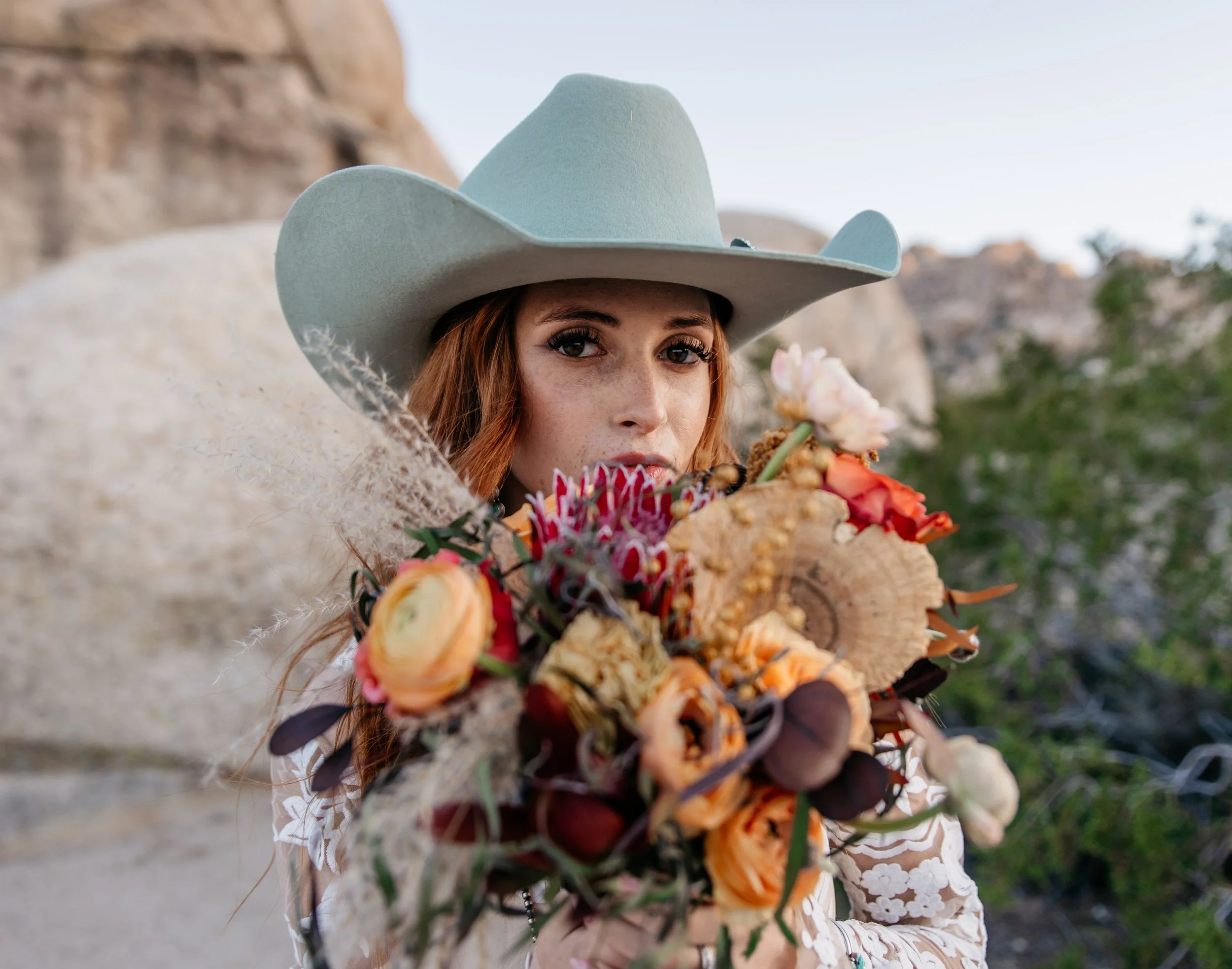 A woman with red hair wearing a light gray cowboy hat holding a bouquet of colorful flowers outdoors with rocky terrain and greenery in the background. timeless Kansas and destination wedding photography