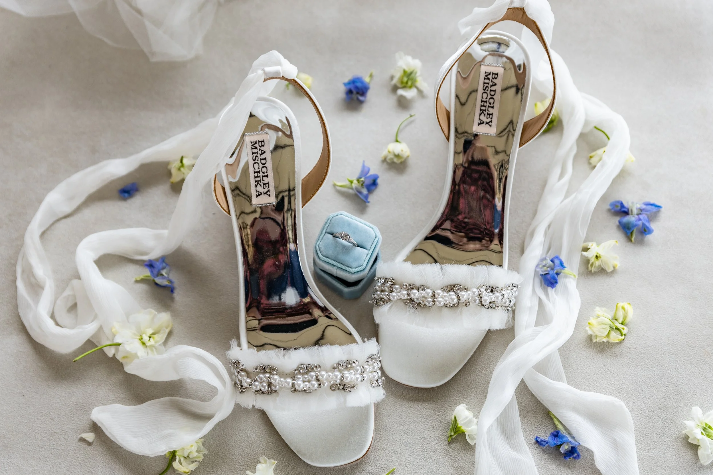 A pair of white high-heeled wedding shoes with pearl and rhinestone embellishments, a light blue ring box with a diamond engagement ring, and a rhinestone bracelet, all surrounded by white and blue flower petals and white ribbon.