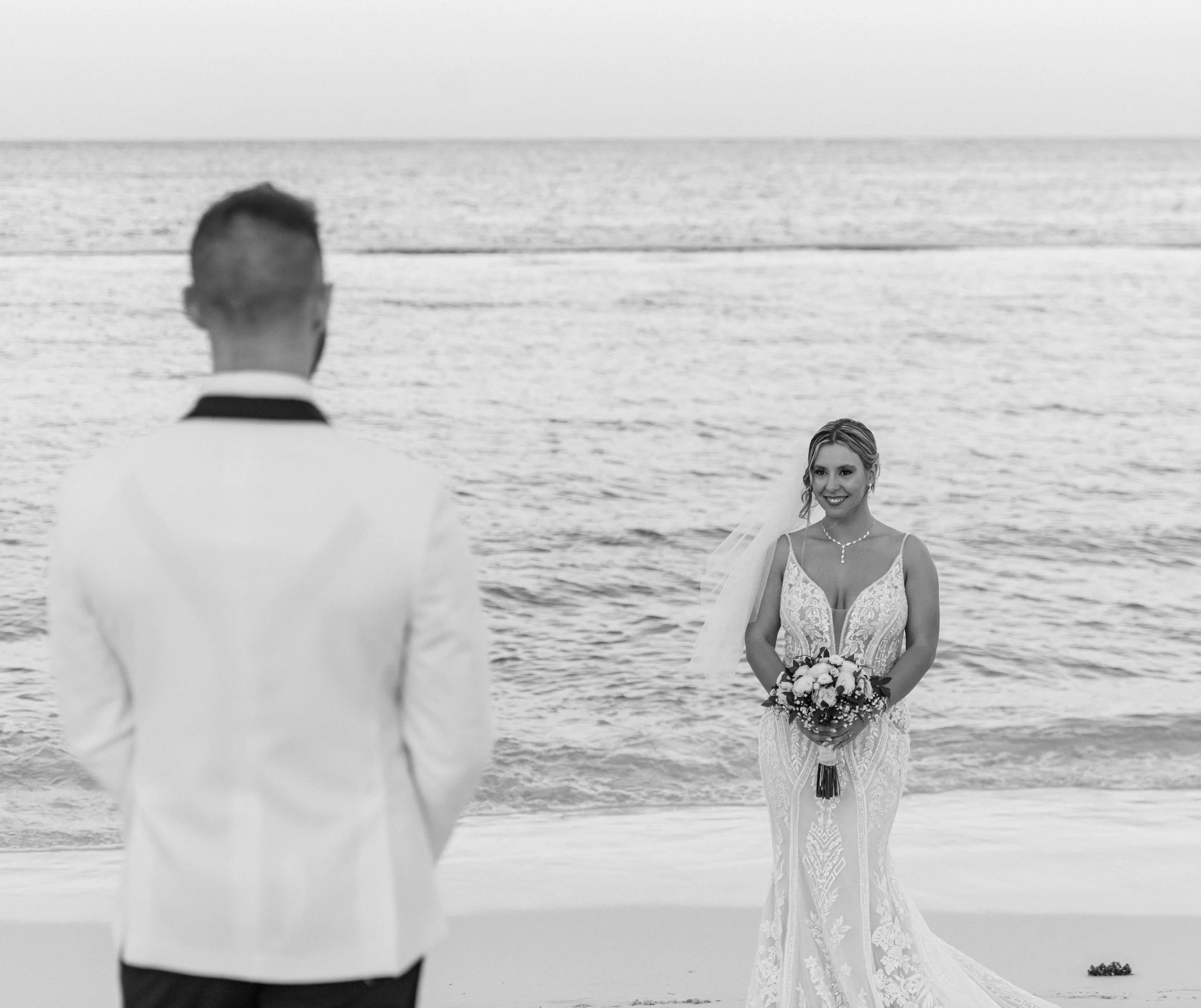 A bride holding a bouquet of flowers gazes at her groom, who is facing her with his back to the camera, on a beach with the ocean in the background. Luxury destination wedding photographer