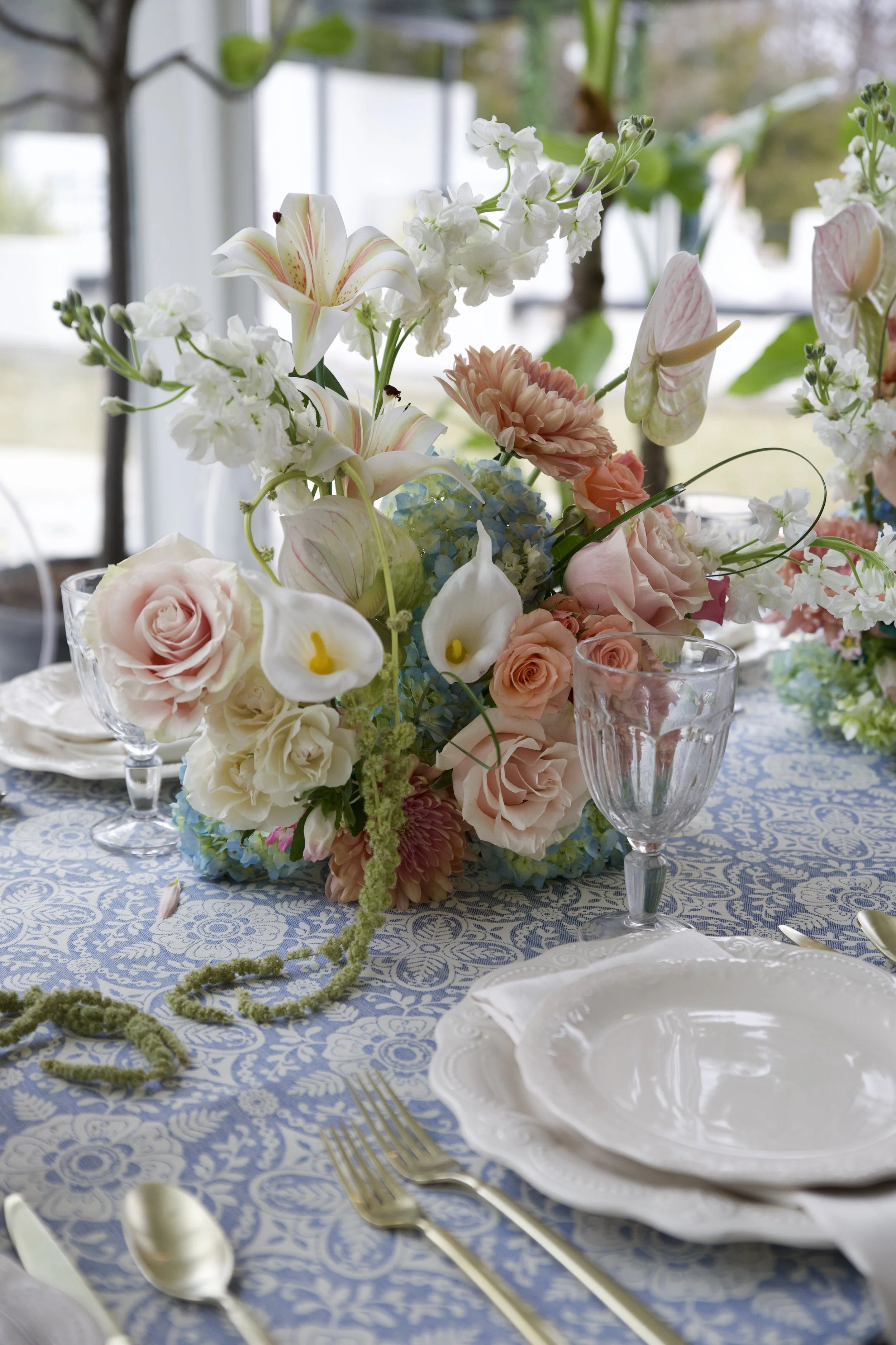 A table setting with a floral centerpiece, white plates, gold cutlery, and glassware on a blue patterned tablecloth. timeless wedding photography