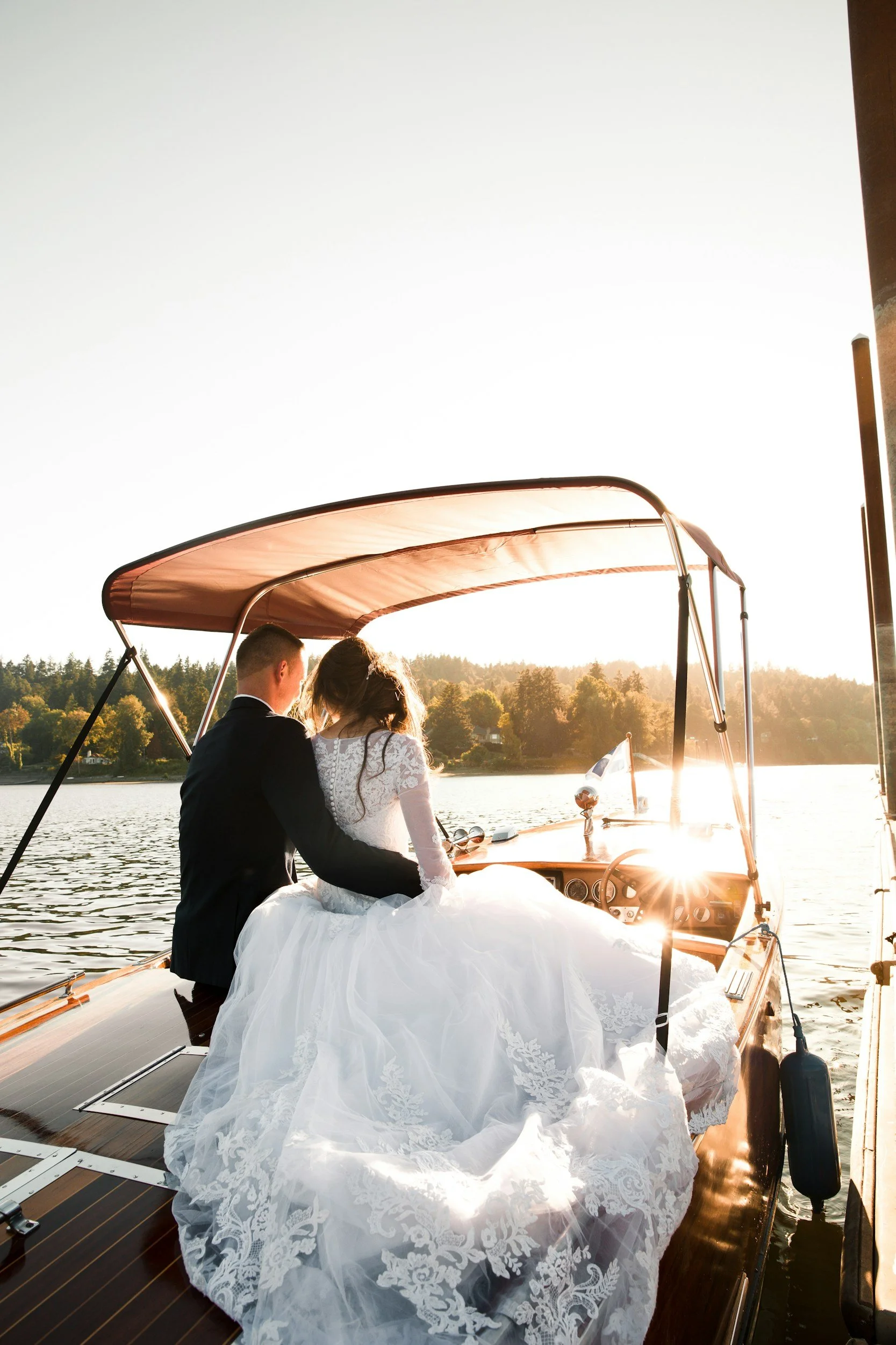 A bride and groom sitting on a boat during sunset, with the bride in a white wedding gown and the groom in a dark suit, on a lake surrounded by trees. Kansas elopement and wedding photography