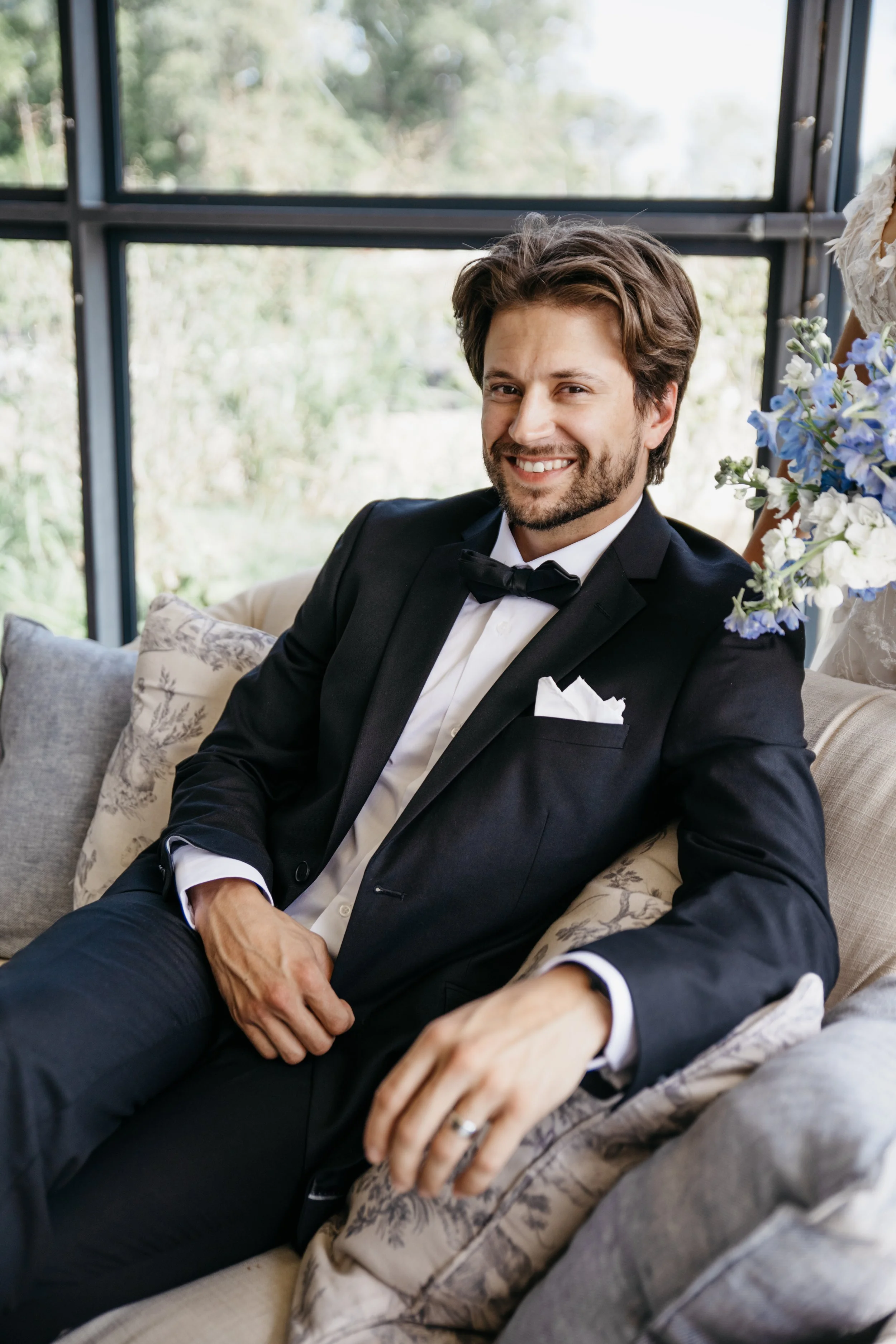 A smiling groom in a tuxedo sitting on a sofa with decorative pillows, holding his hand on his lap, with a large window and greenery in the background.  Luxury wedding photography in Kansas and worldwide