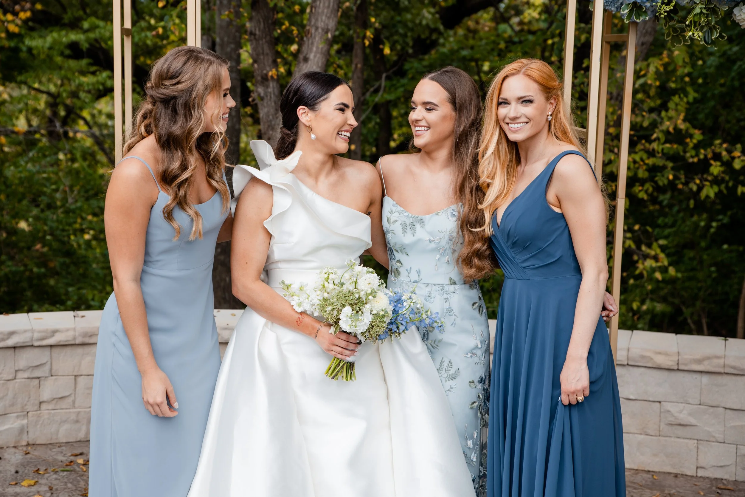 A bride and three bridesmaids posing outdoors in front of trees and a stone wall, smiling and laughing. Kansas wedding photographer
