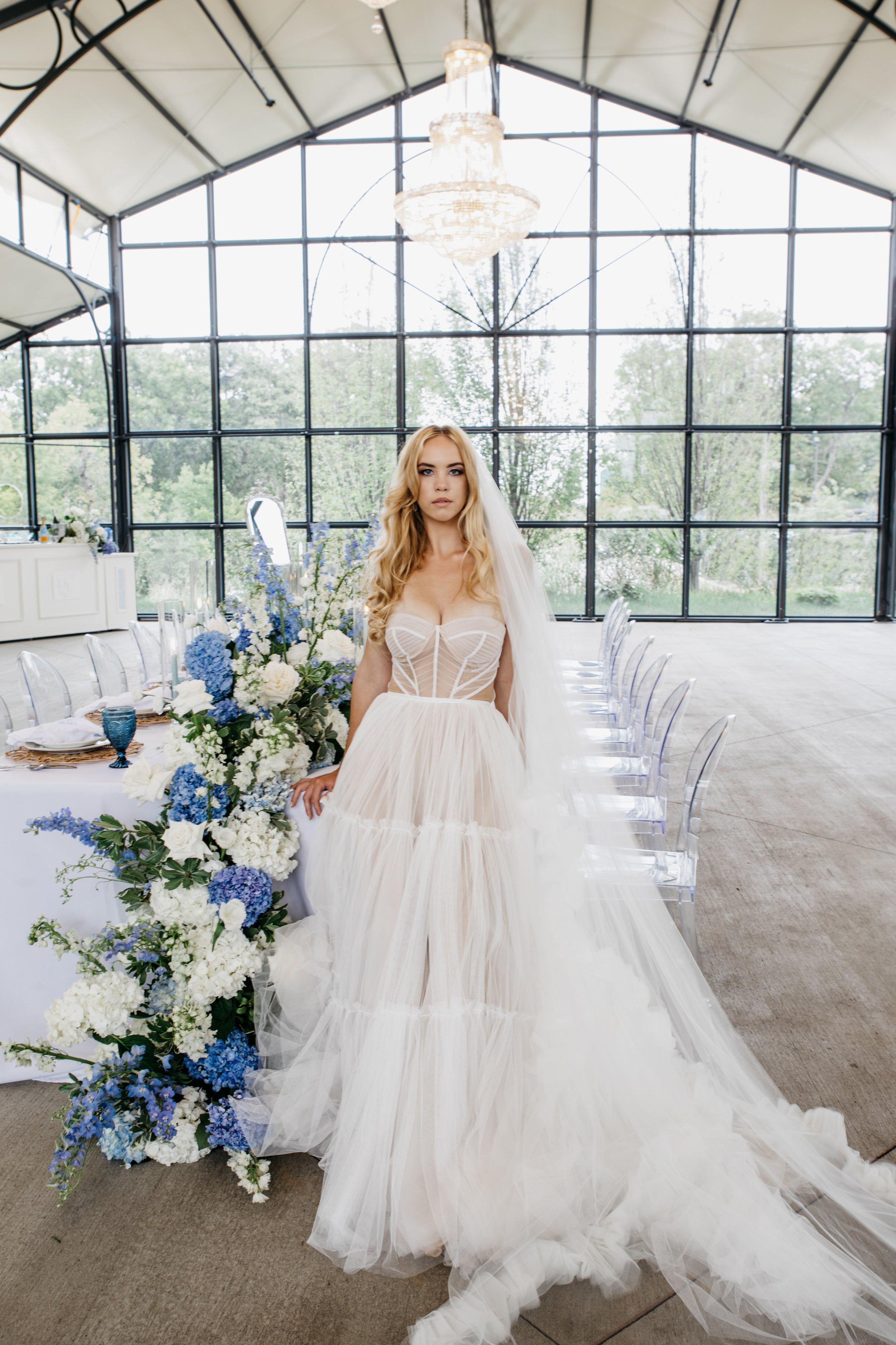 Bride in a white wedding dress standing next to a large floral arrangement in a bright, glass-wall wedding venue. International tieless elopement and wedding photographer