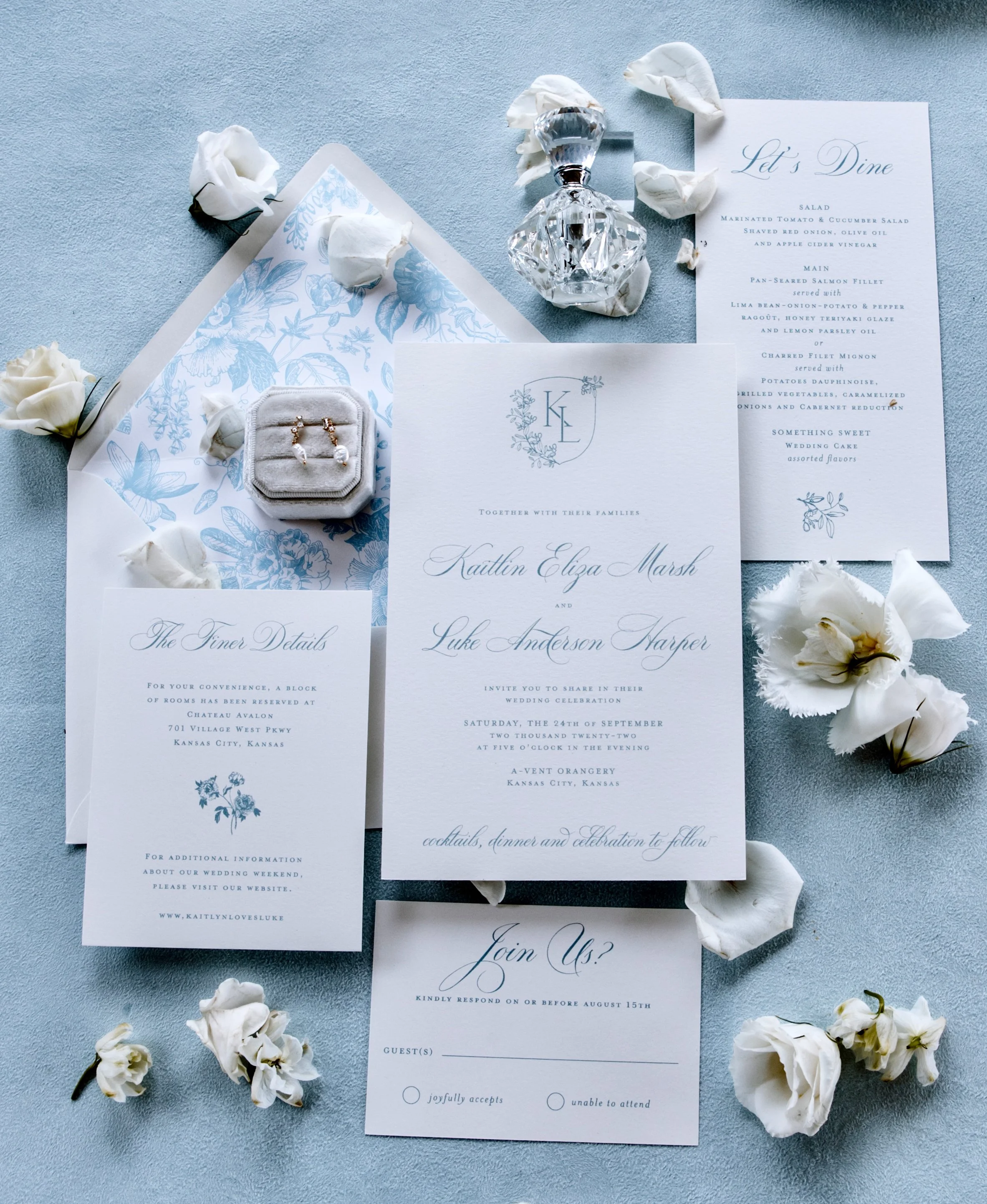 Wedding invitation suite with rings, a perfume bottle, and white floral accents on a light blue surface.  Luxury wedding photography in Kansas City and all over the world