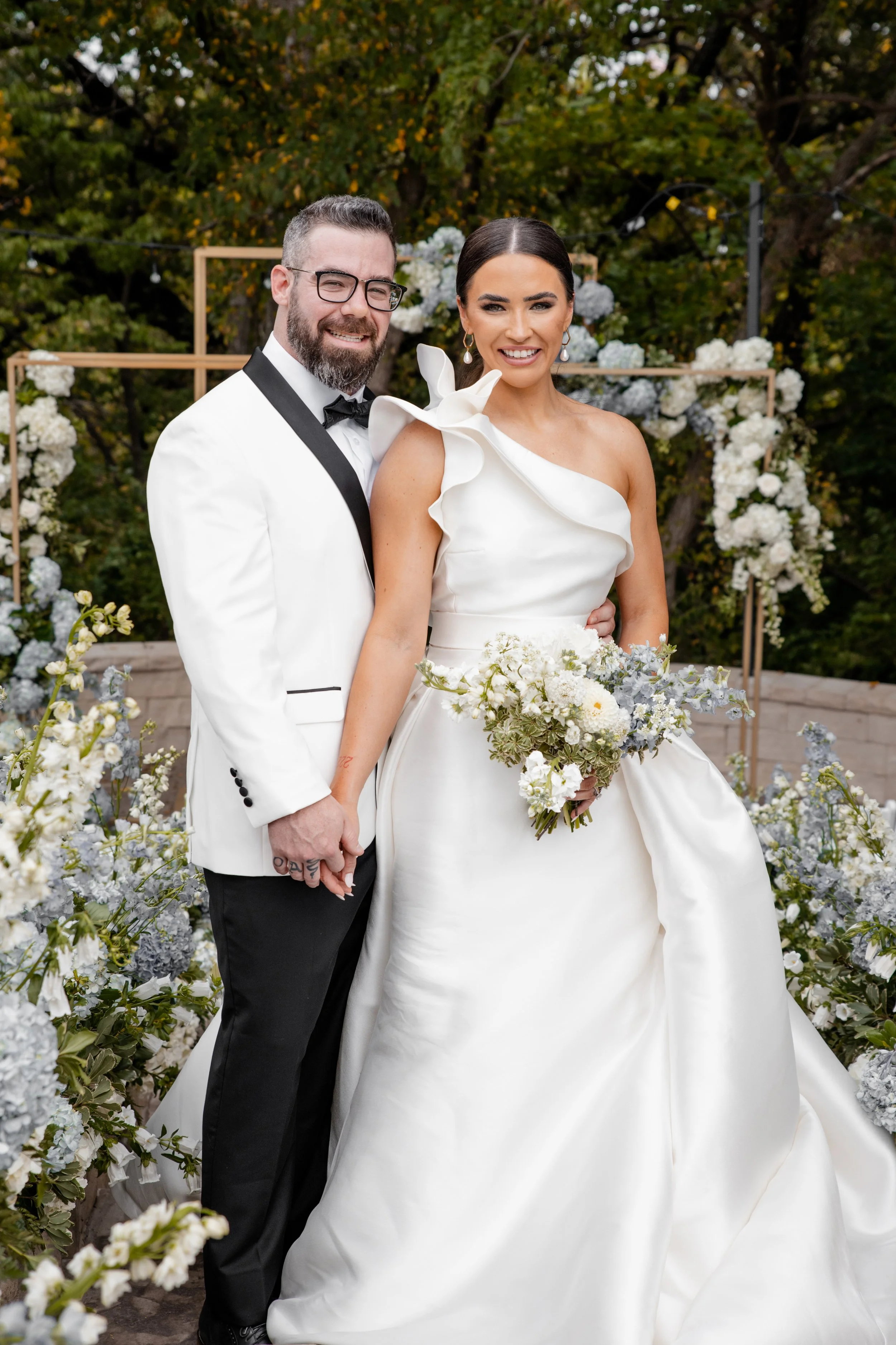 A newlywed couple standing outdoors, holding hands, amidst white and gray floral decorations, with trees in the background. The groom wears a white tuxedo jacket with black lapels, black pants, and glasses. The bride wears a white one-shoulder wedding gown with ruffles, holding a bouquet of white and gray flowers, with dark hair styled in an updo.