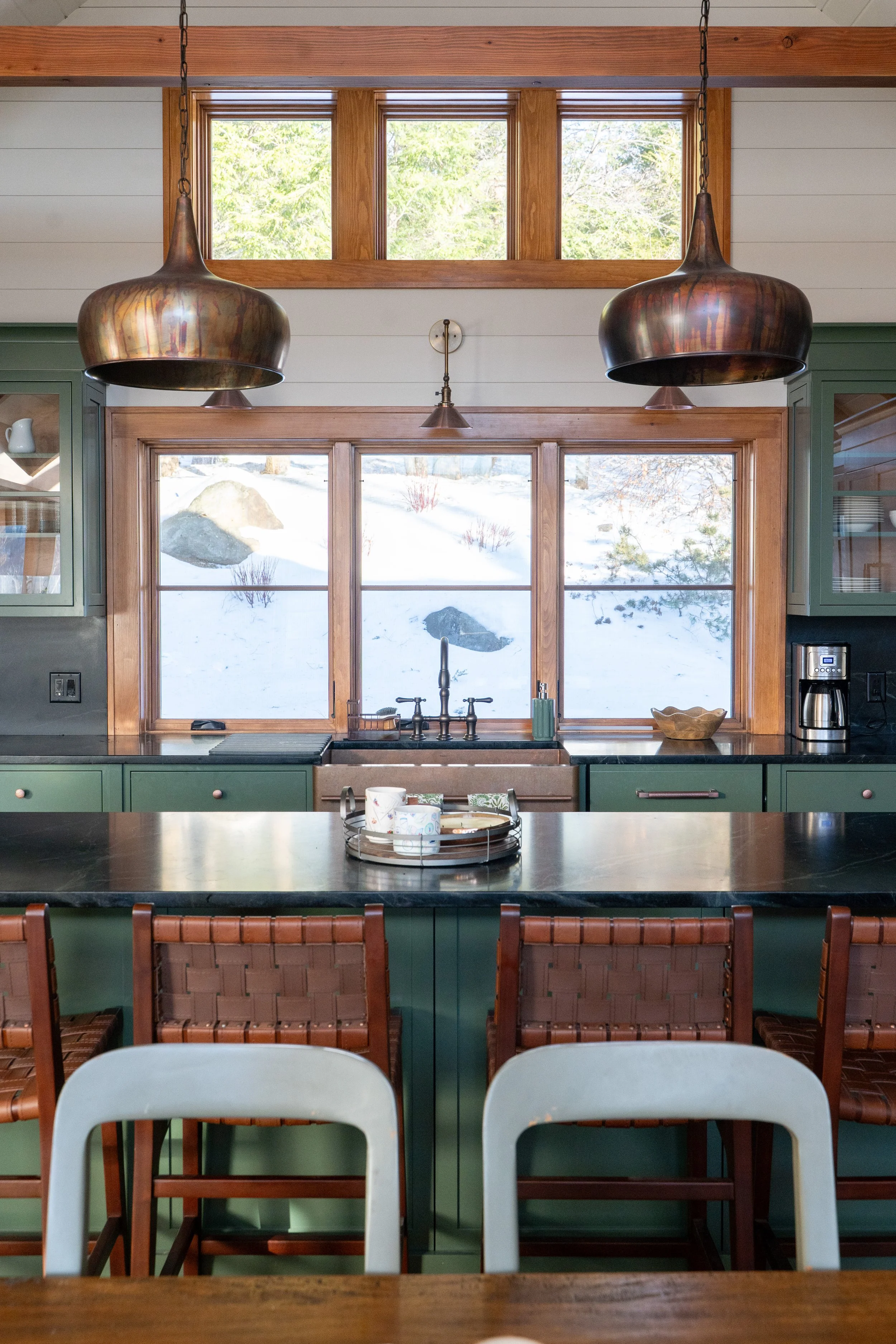 Kitchen with large window showing snowy landscape outside, green cabinets, black countertop, and three woven chairs in front.