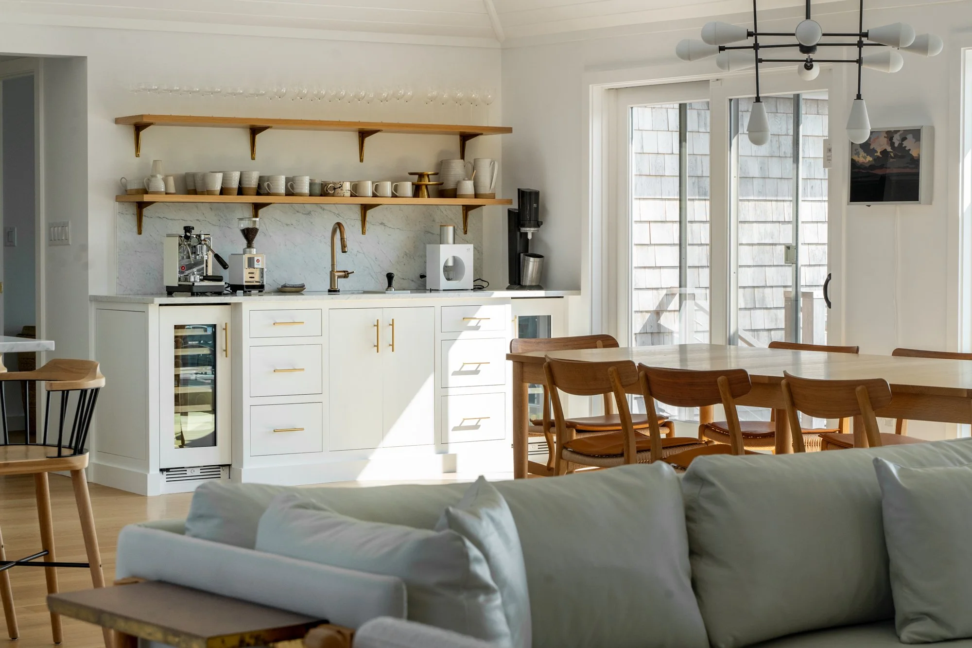 Open-concept kitchen with white cabinets, wooden open shelves, and a marble backsplash. A coffee machine and espresso machine are on the counter. A wooden dining table with matching chairs is nearby, and large glass sliding doors lead to a balcony. A