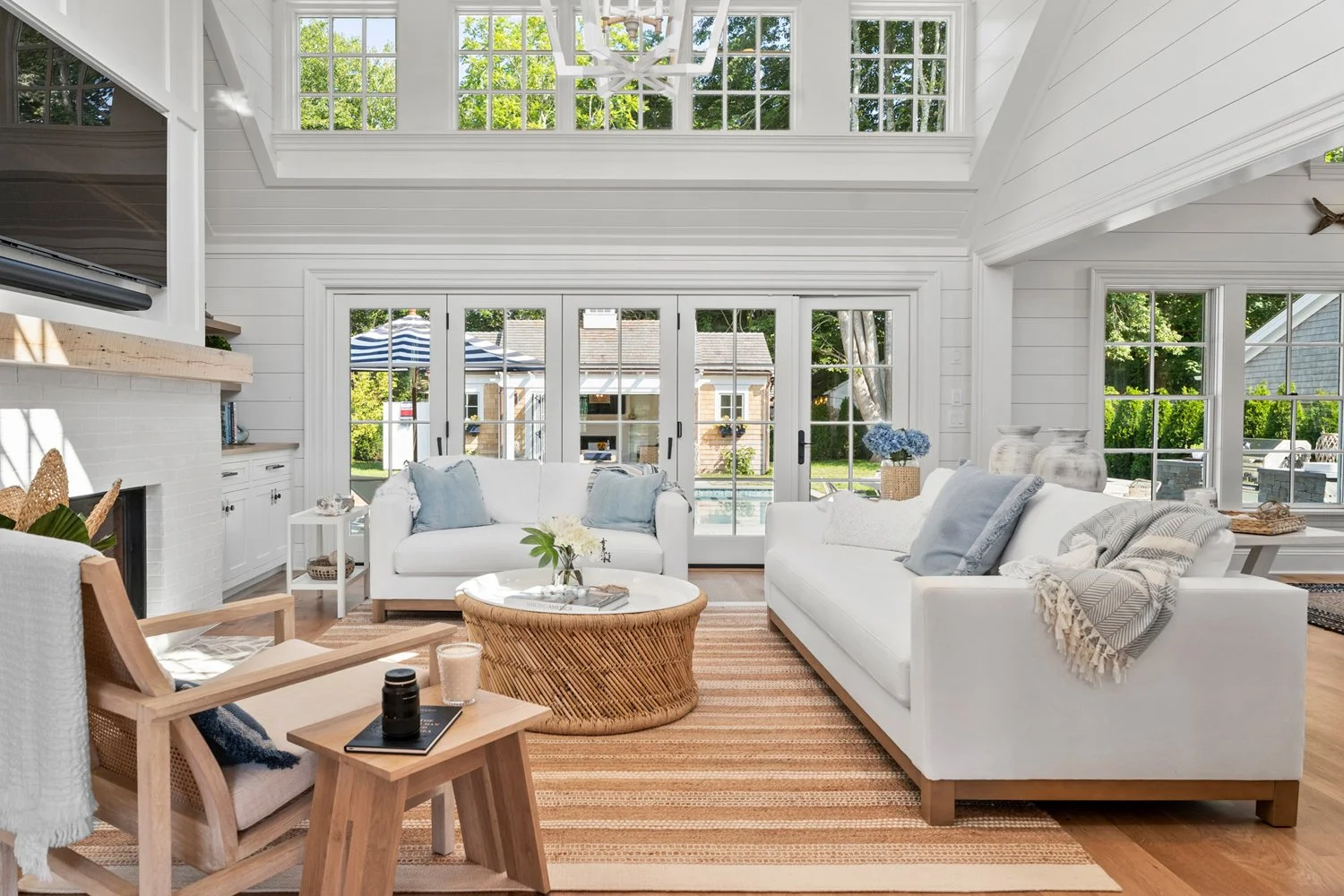 Bright living room with white sofas and armchair, glass doors leading outside to a backyard with a pool, wooden coffee table with flowers, striped rug, and large windows allowing natural light.