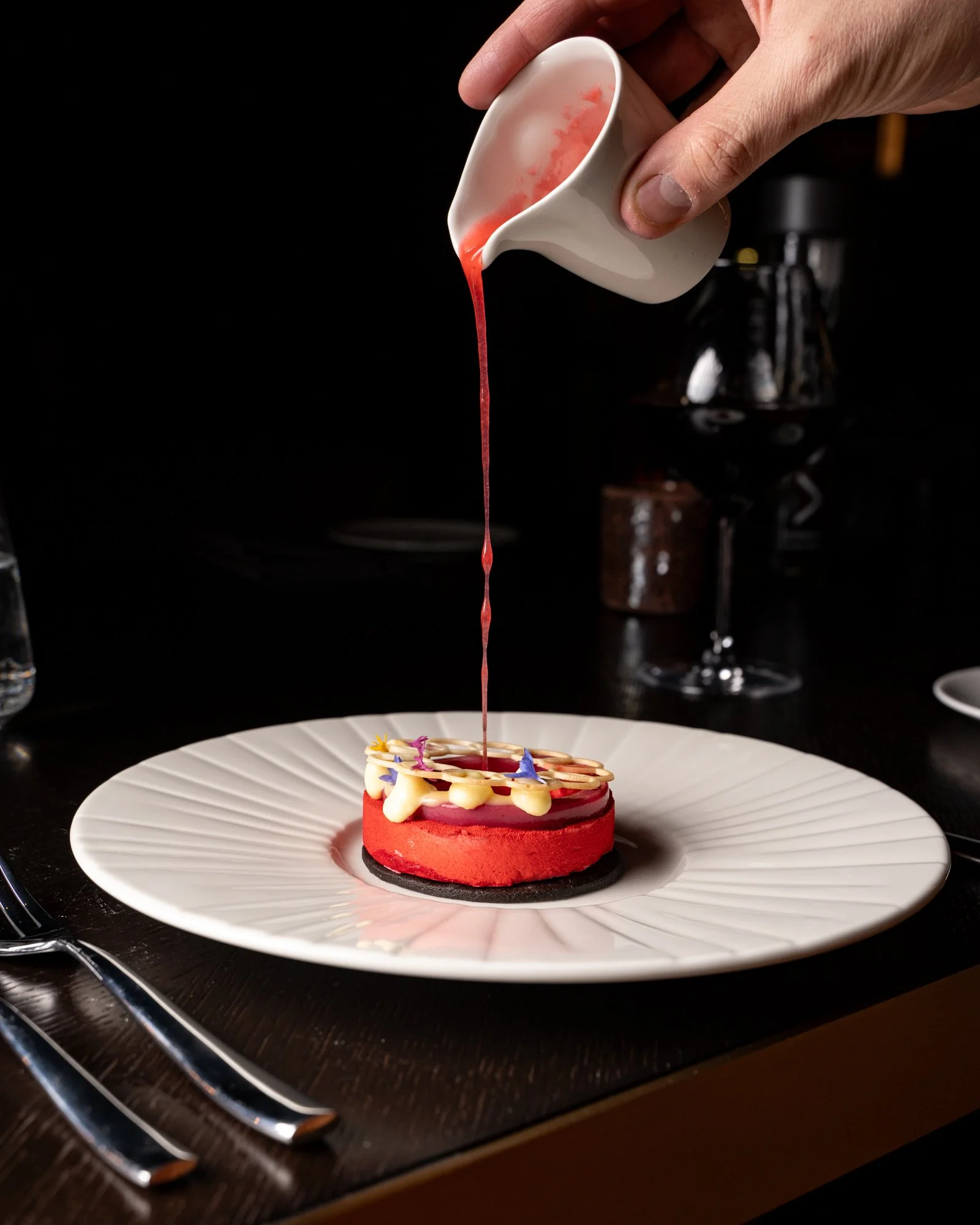 A dessert on a white plate with a black base, decorated with colorful edible flowers and white chocolate, is being topped with pink sauce from a small white pitcher held by a hand, against a black background.