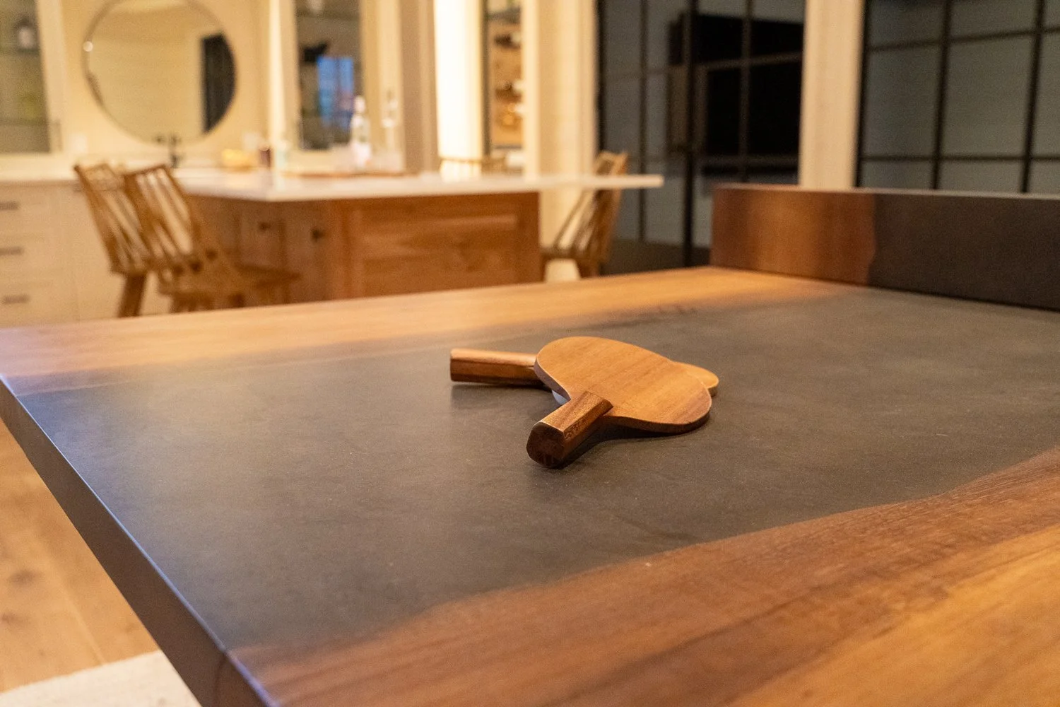 A wooden table with a small wooden paddle-shaped object on a dark mat surface in a cozy kitchen.