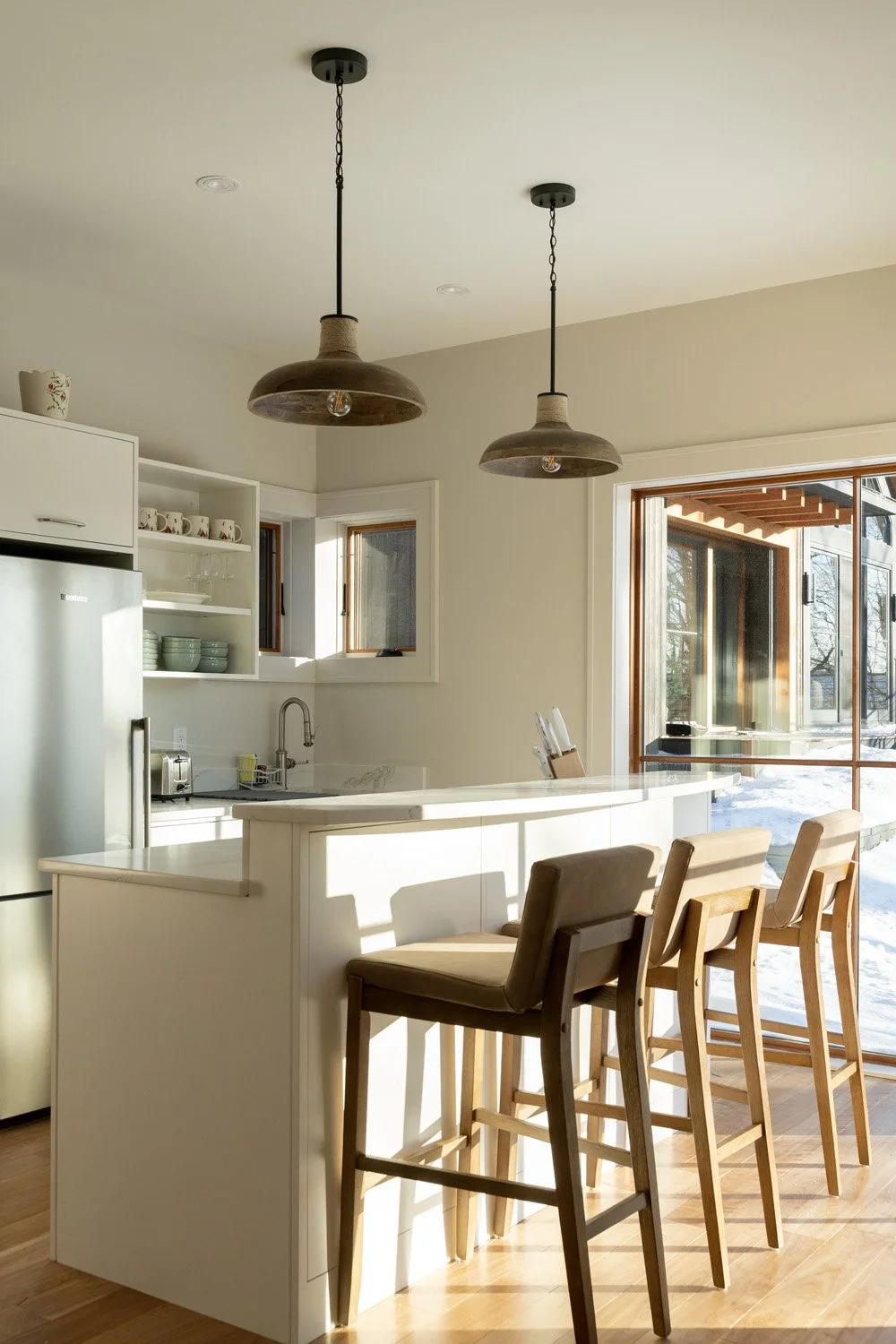 Bright kitchen with white island, brown bar stools, hanging pendant lights, open cabinets, stove, sink, and large glass door showing outdoor snow scene.