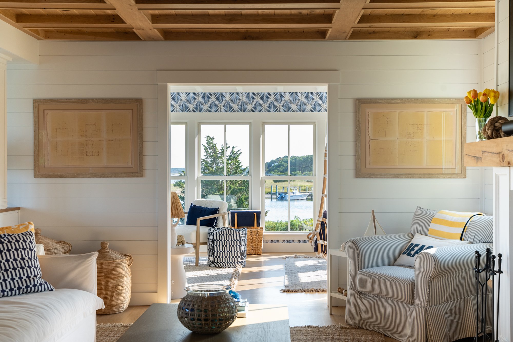 A cozy living room with white shiplap walls and a natural wood ceiling. There's a striped white and gray armchair, a white couch with navy and white pillows, a wicker basket, and a coffee table with a decorative vase. Large windows at the back show a view of a waterway with boats and green landscape.
