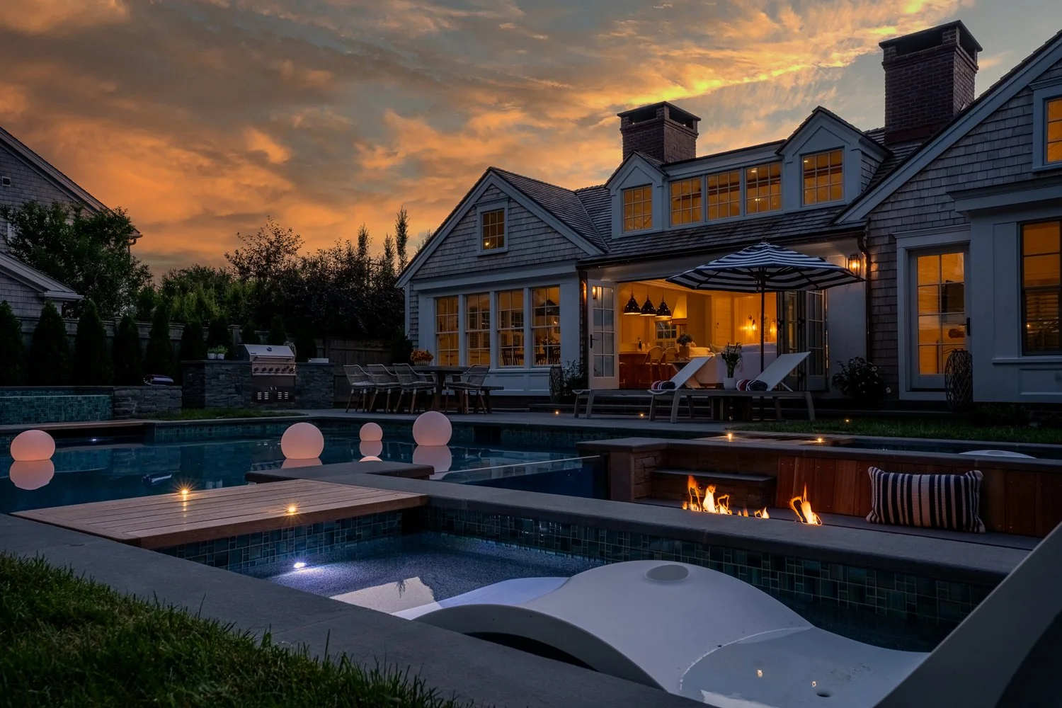 A backyard with a swimming pool, illuminated by floating orbs and a fire feature, adjacent to a large house with lit windows and outdoor seating, during sunset with a colorful sky.