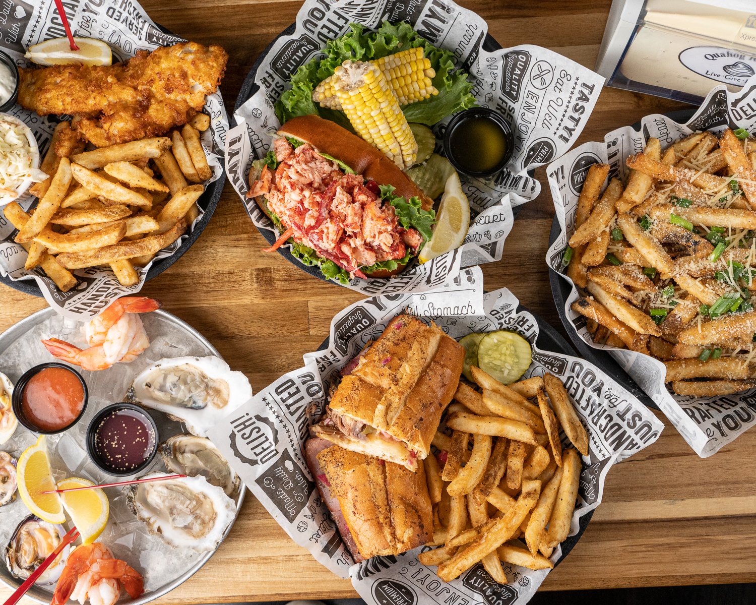 Assorted seafood and fried dishes on a wooden table, including fried fish with fries, lobster roll with fries, shrimp and oysters with dipping sauces, and pasta with grated cheese and green onions.