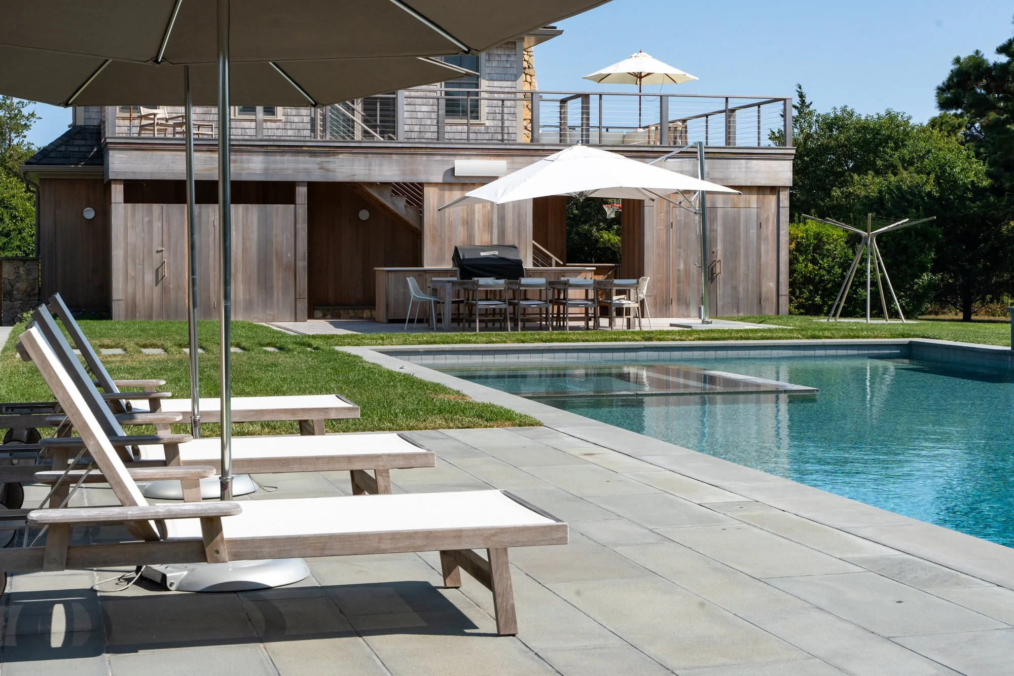 Backyard with swimming pool, wooden deck, outdoor dining table with chairs, lounge chairs, umbrellas, and a wooden house with balcony.