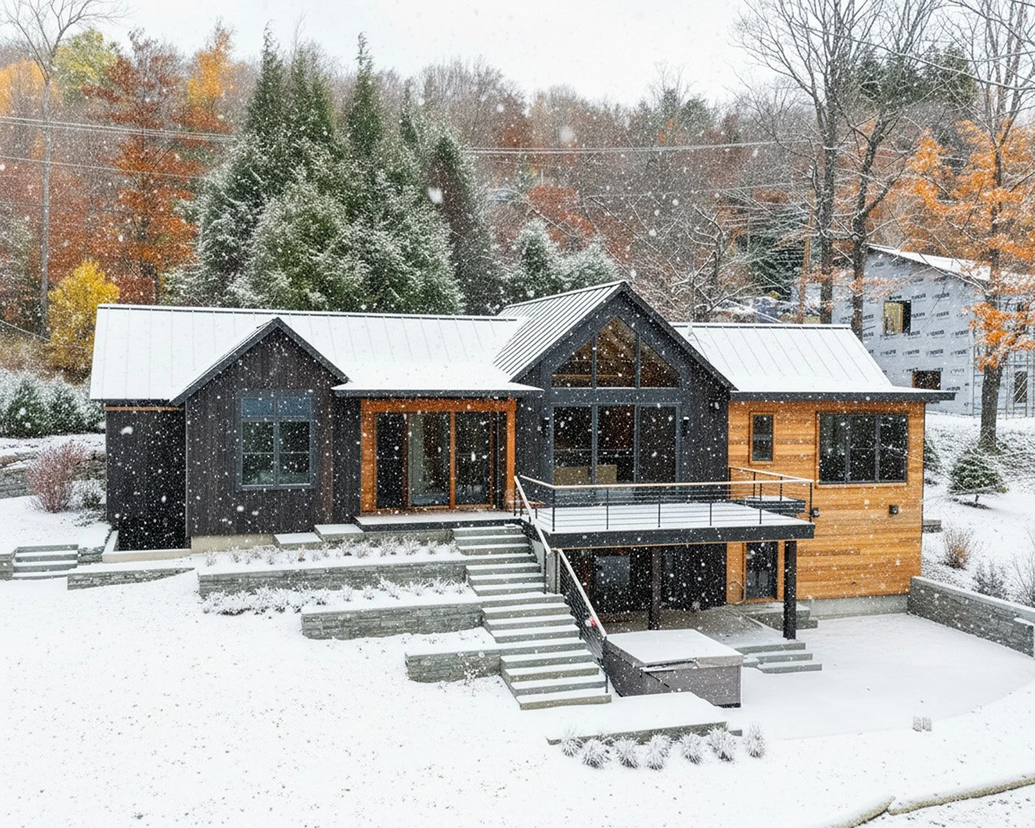 Modern house with black and wooden exterior, surrounded by snow and falling snowflakes, with trees displaying fall colors in the background.