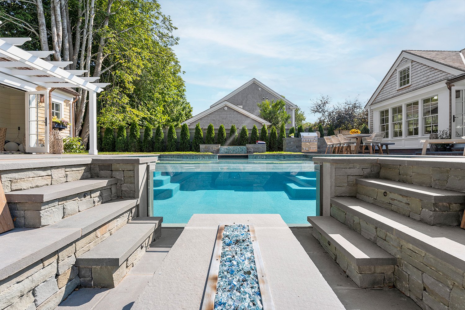 Backyard with a swimming pool, stone steps, and a landscaped patio area. There is outdoor furniture, a grill, and a hedgerow fence with houses and trees in the background.