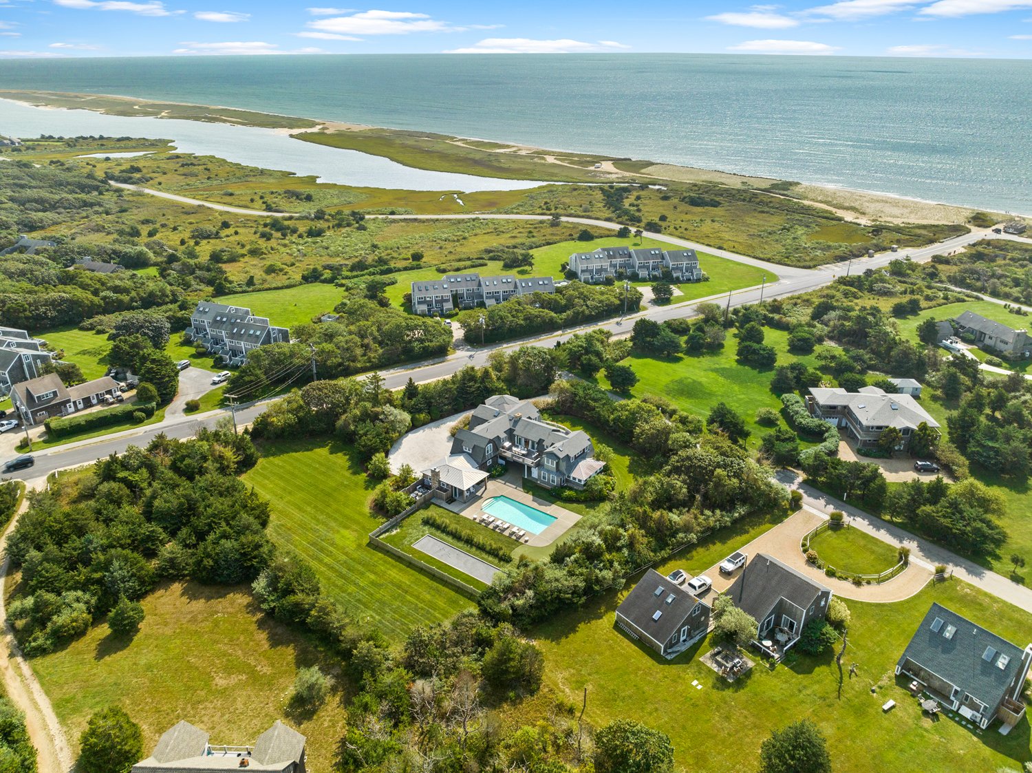 Aerial view of a coastal neighborhood with houses, green lawns, a swimming pool, and a shoreline with a body of water in the background.