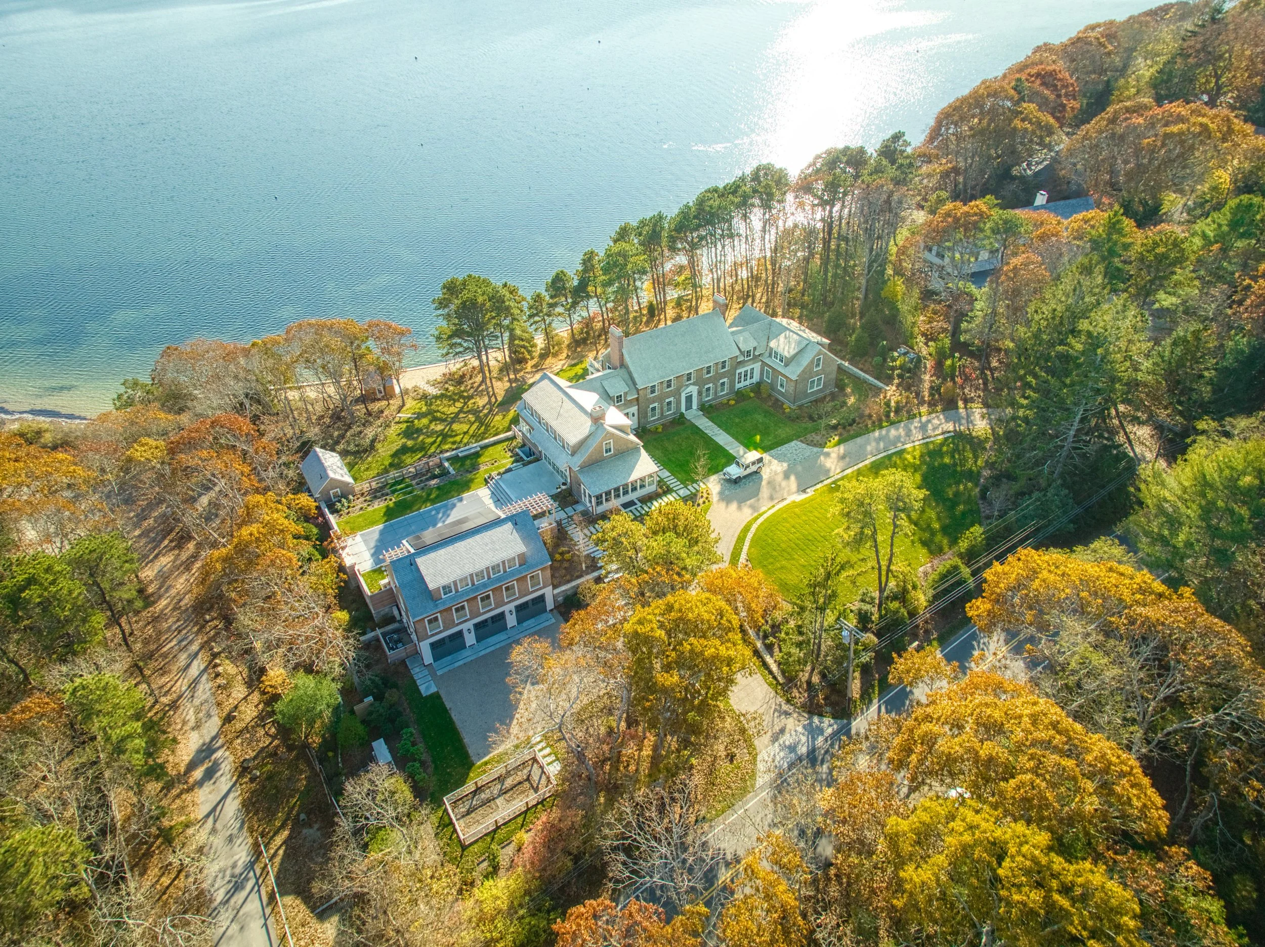 Aerial view of a large house with a gray roof next to a lake, surrounded by colorful autumn trees and a driveway winding through the property.