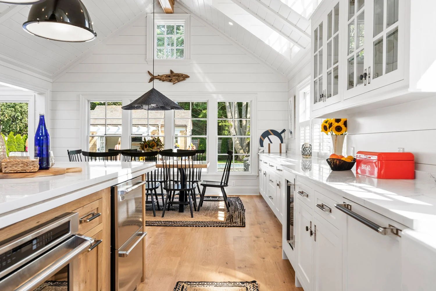 Bright kitchen and dining area with white shiplap walls, large windows, black chairs around a dinner table, and wooden flooring.