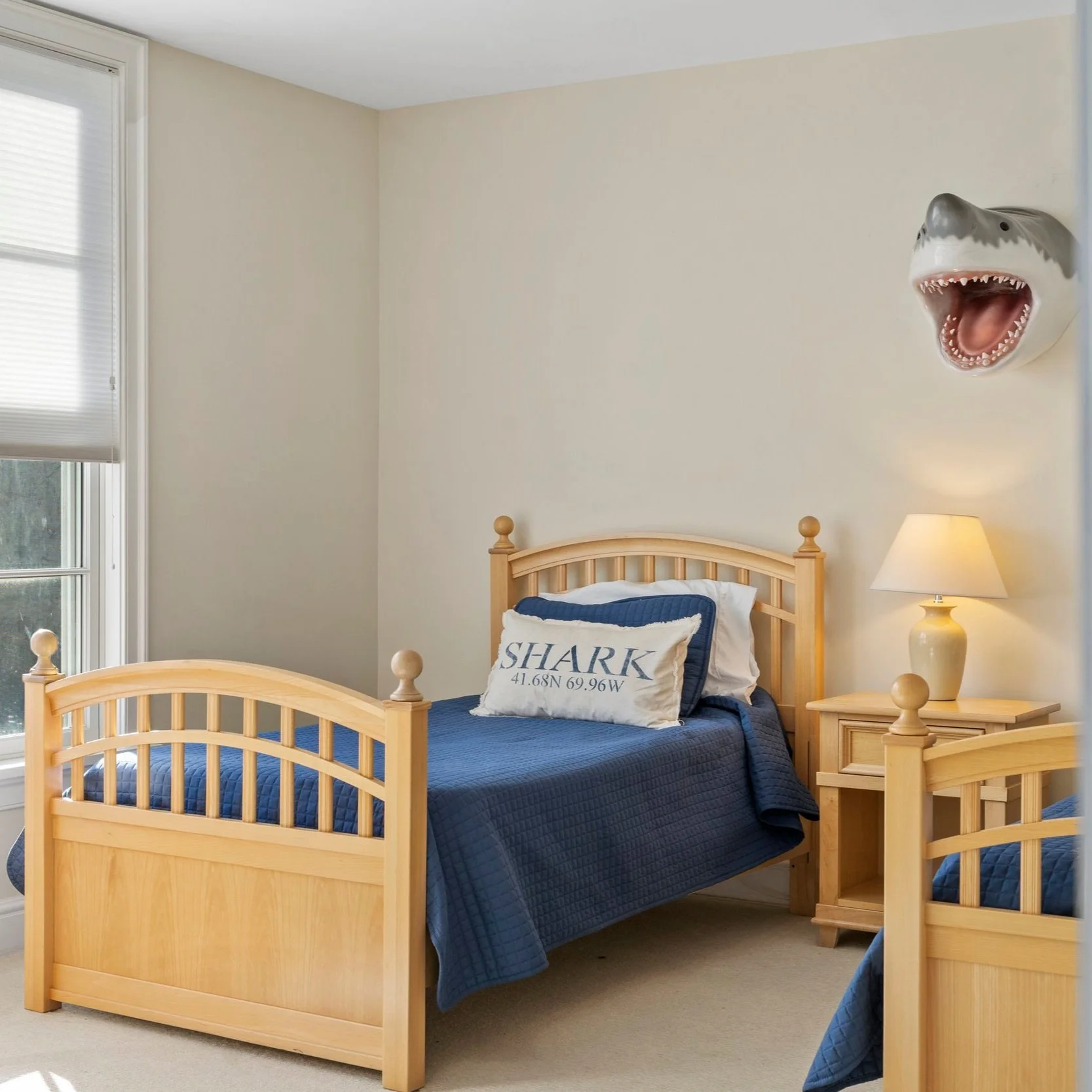 A bedroom with two single beds made of light wood, each with blue quilted bedspreads and pillows. There is a window with white blinds on the left wall, and a nightstand with a lamp on the right. A shark head wall decoration is mounted on the wall above the beds.