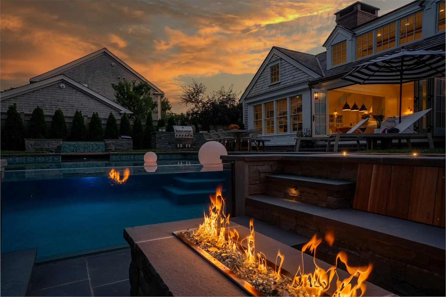 A backyard view at sunset featuring a swimming pool with floating lights, a fire feature, lounge chairs, a patio with an umbrella, and a large house with illuminated windows.