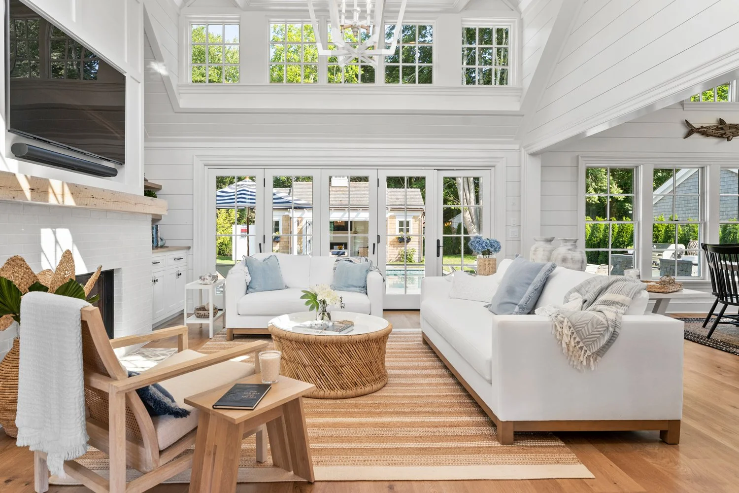 Bright living room with white walls, large windows, and sliding glass doors leading outside. Contains white sofas with light blue and gray pillows, a round wicker coffee table, a wooden armchair with a white blanket, and a striped area rug on wooden floors. Outside features a patio with a striped umbrella and backyard greenery.