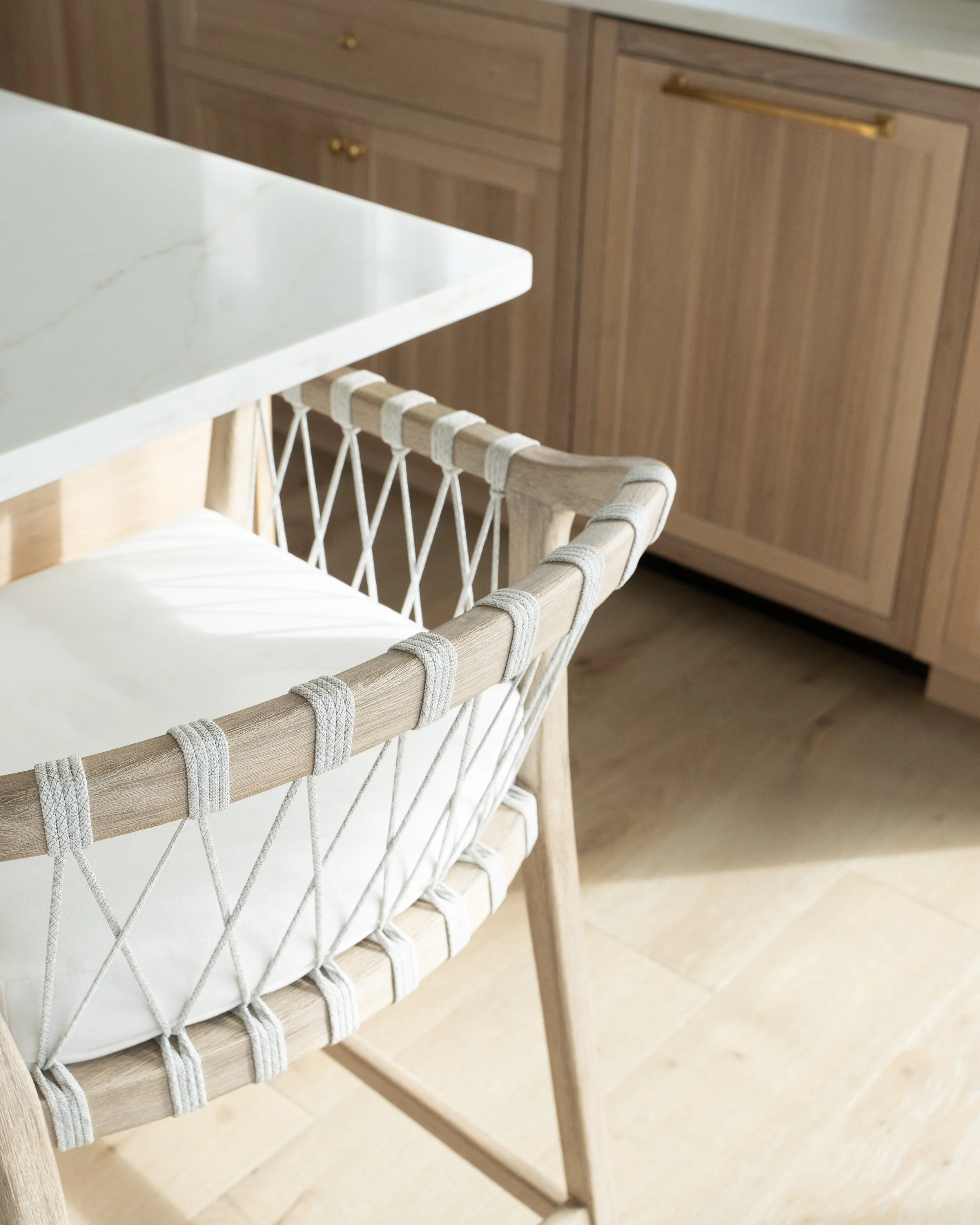 Close-up of a wooden chair with a woven rope backrest and white cushion, next to a white marble-top table, in a kitchen with wooden cabinets.