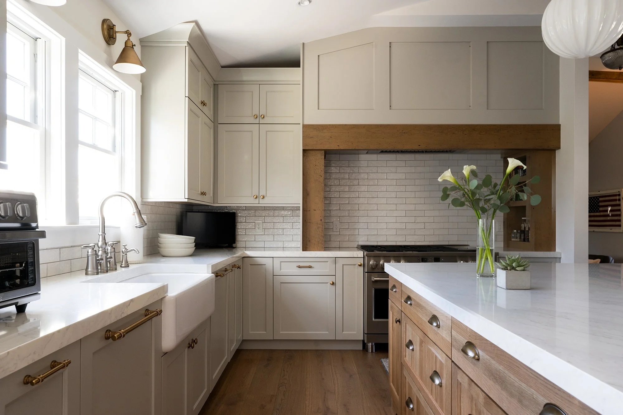 Modern kitchen with white cabinets, a farmhouse sink, marble countertops, and a mixed wood island. There are large windows, a small microwave, and a vase with flowers on the island.