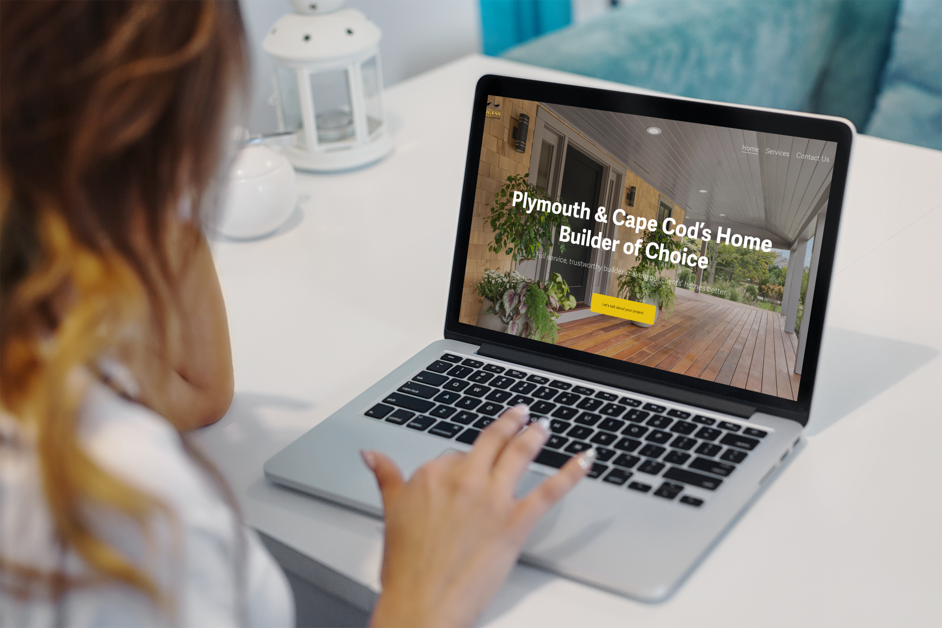 A woman with long hair working on a laptop displaying a website for a home builder service, with a porch and plants visible in the background.