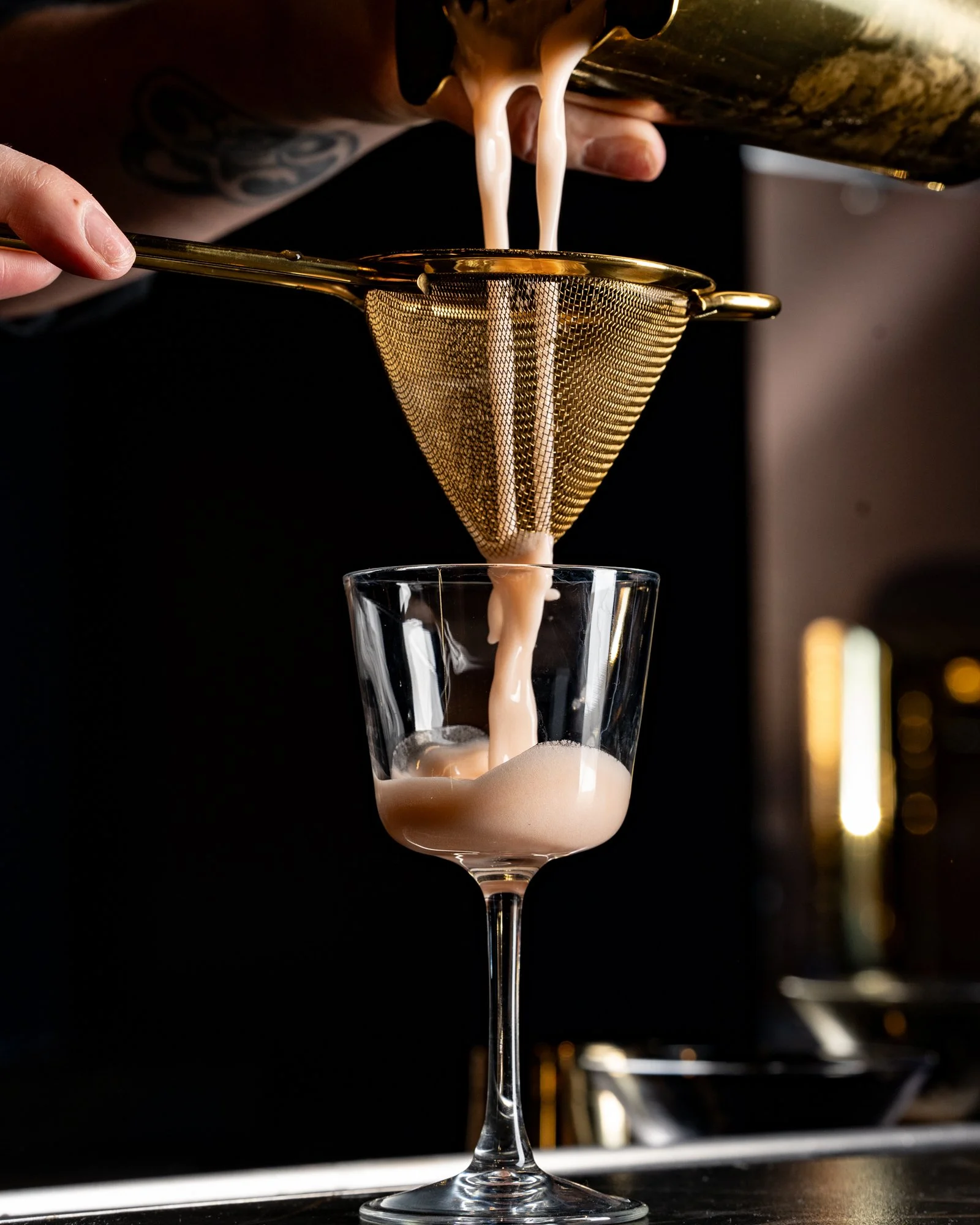 A bartender pours a creamy cocktail through a gold strainer into a cocktail glass.