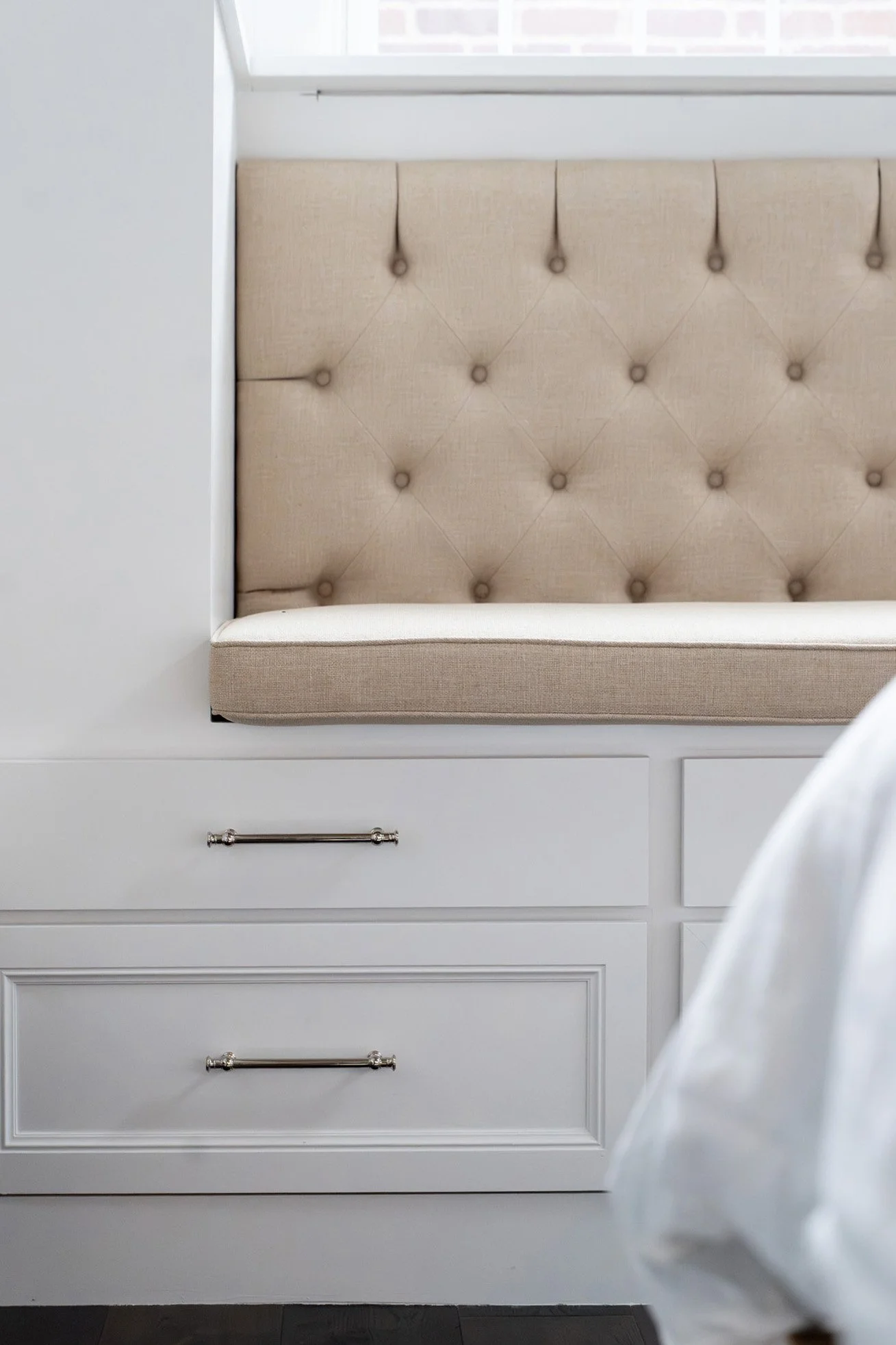 A built-in window seat with a beige, tufted, fabric-backrest cushion, integrated into white cabinetry with chrome handles.