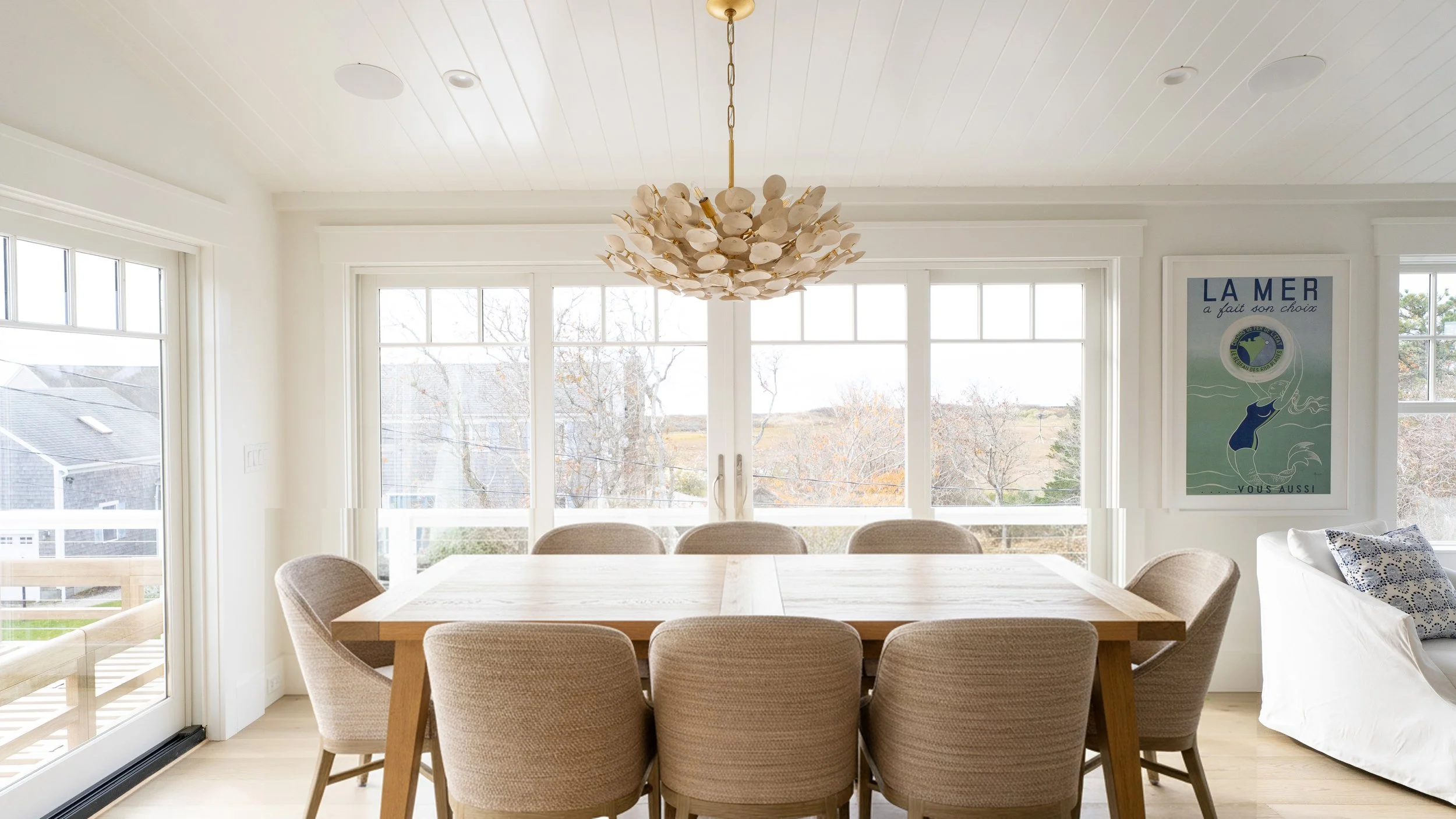 Bright dining room with a wooden table and eight beige upholstered chairs, large windows with a view of trees and houses outside, a white sofa, and a hallway with large glass sliding doors.