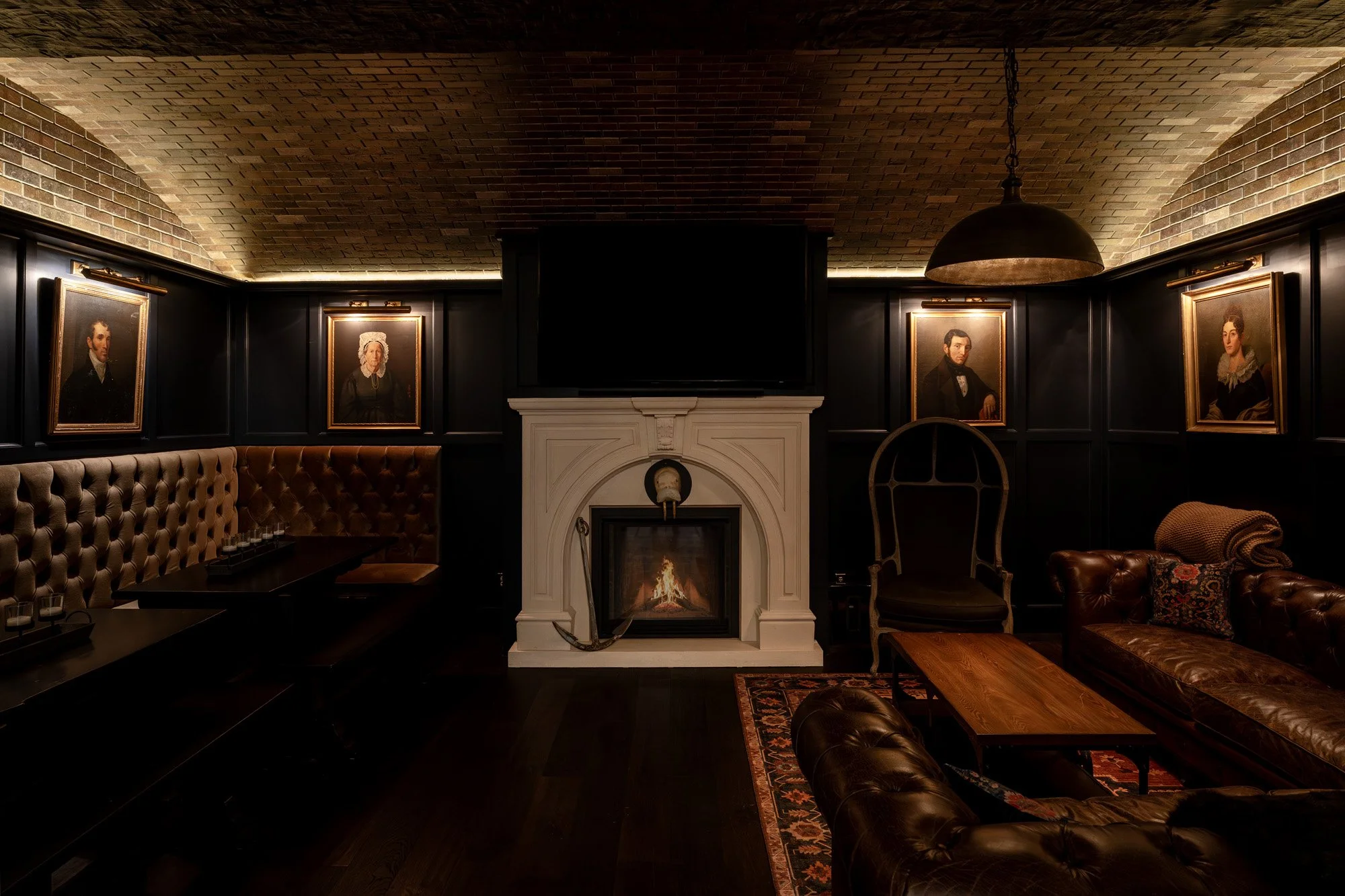 A cozy, dimly lit lounge with a brick vaulted ceiling, dark paneled walls decorated with portrait paintings, a white fireplace with a mirror and clock above, leather sofas, and a large hanging lamp.