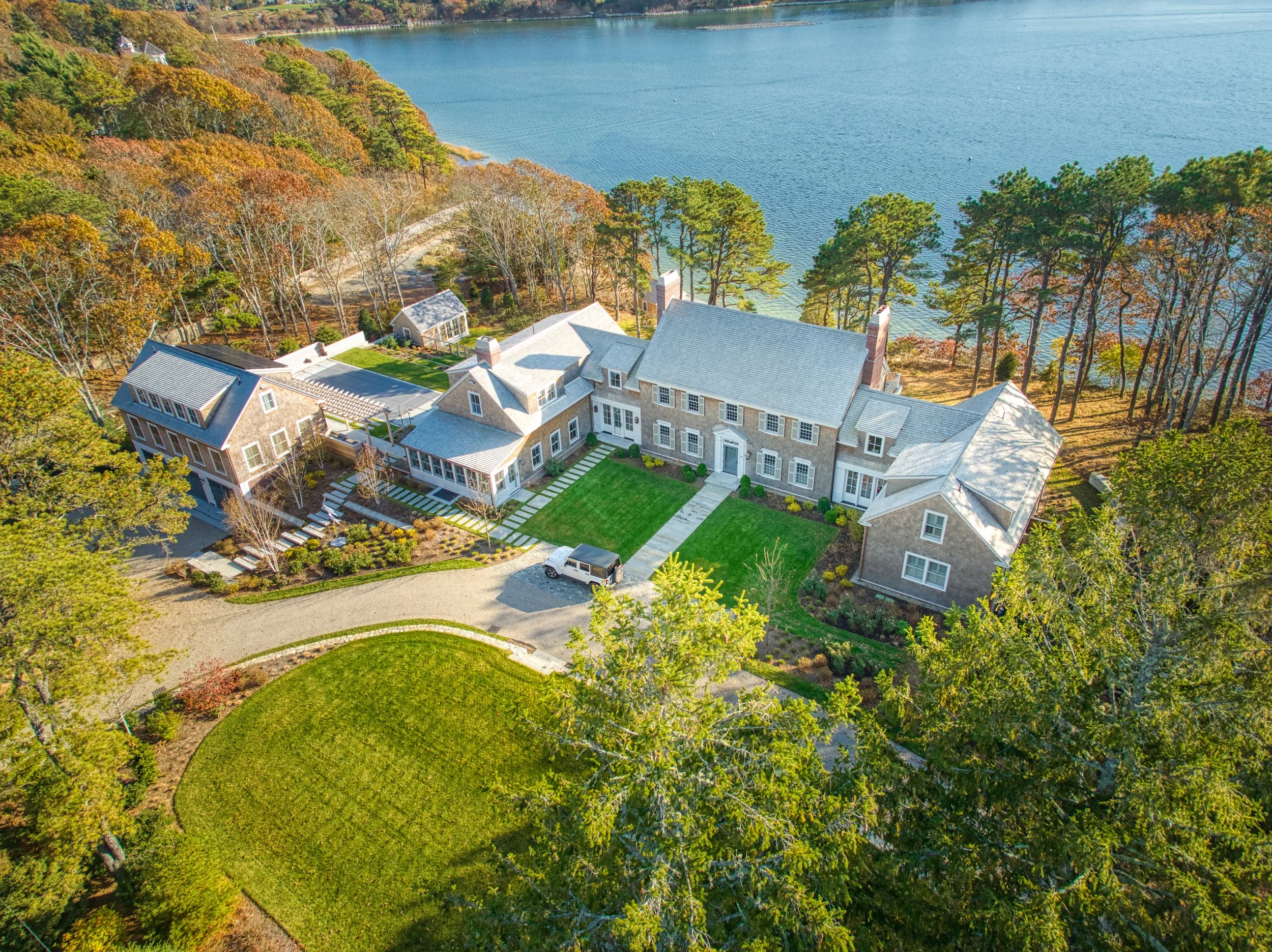 A large house with gray exterior, surrounded by trees and a grassy yard, lakeside view in the background.