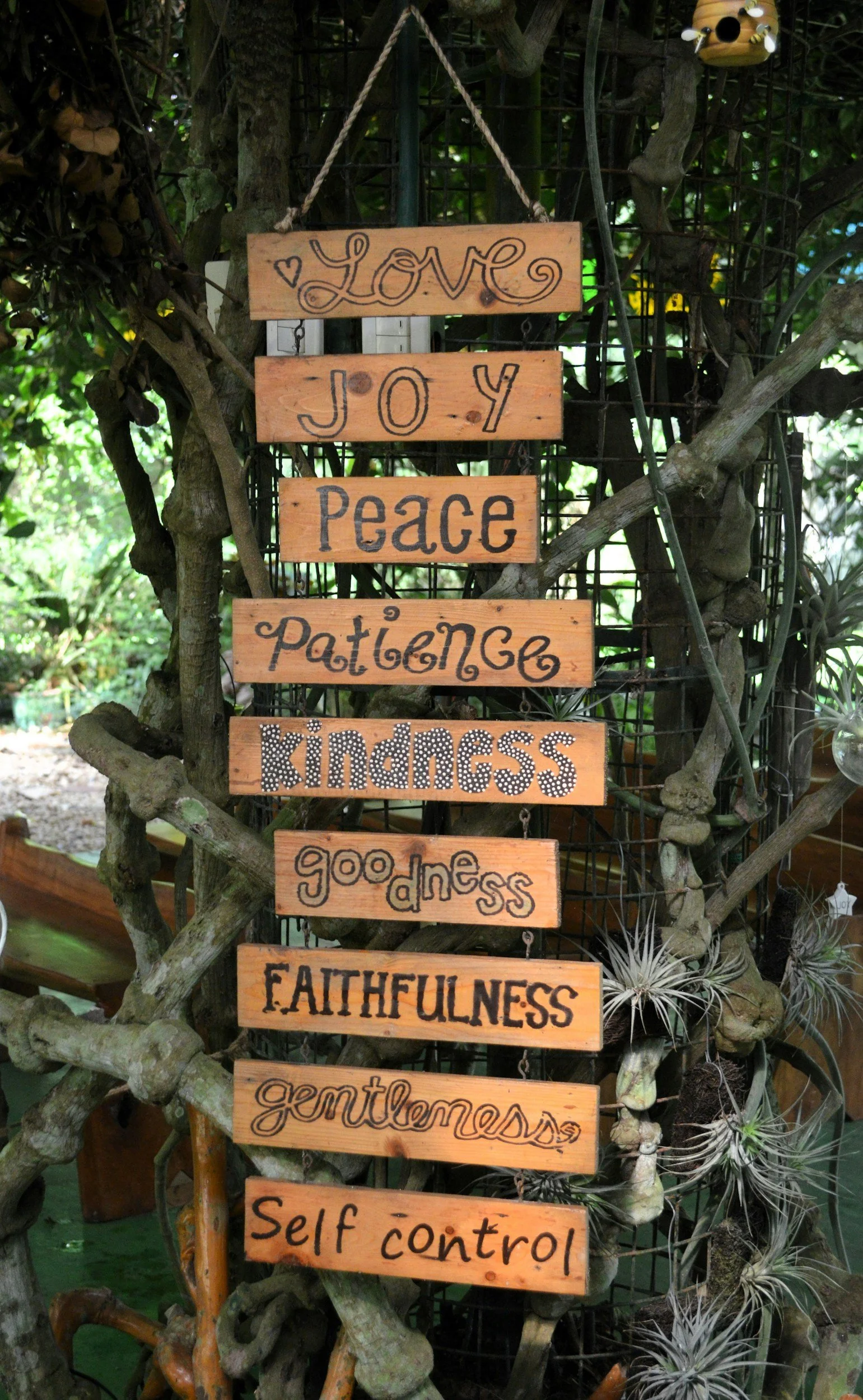 A wooden sign with multiple segments hanging on a vine-covered trellis, displaying words such as 'Love,' 'Joy,' 'Peace,' 'Patience,' 'Kindness,' 'Goodness,' 'Faithfulness,' 'Gentleness,' and 'Self Control' in decorative fonts.