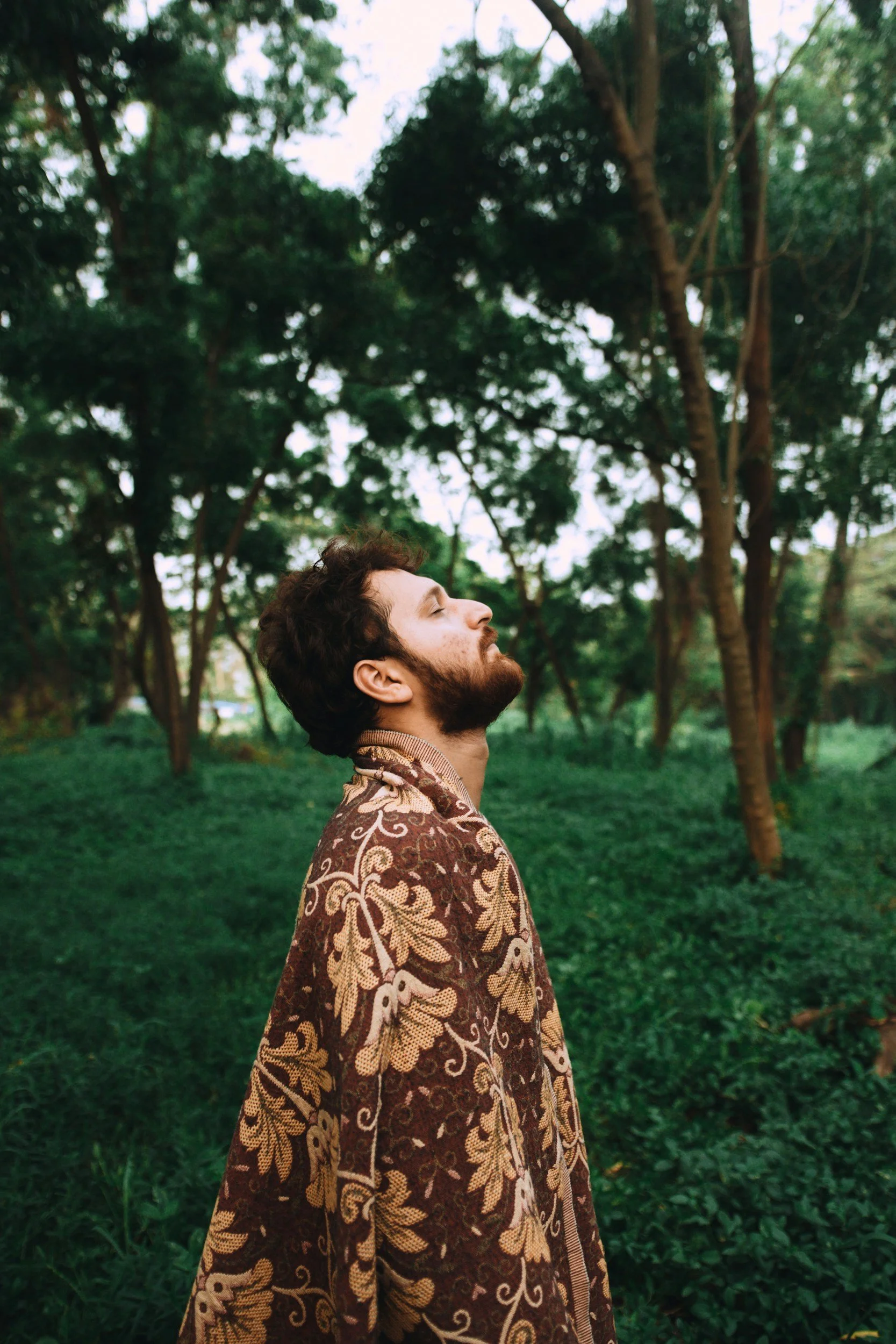 A man with closed eyes and beard facing up standing in a green park with trees in the background, wearing a brown patterned shirt.