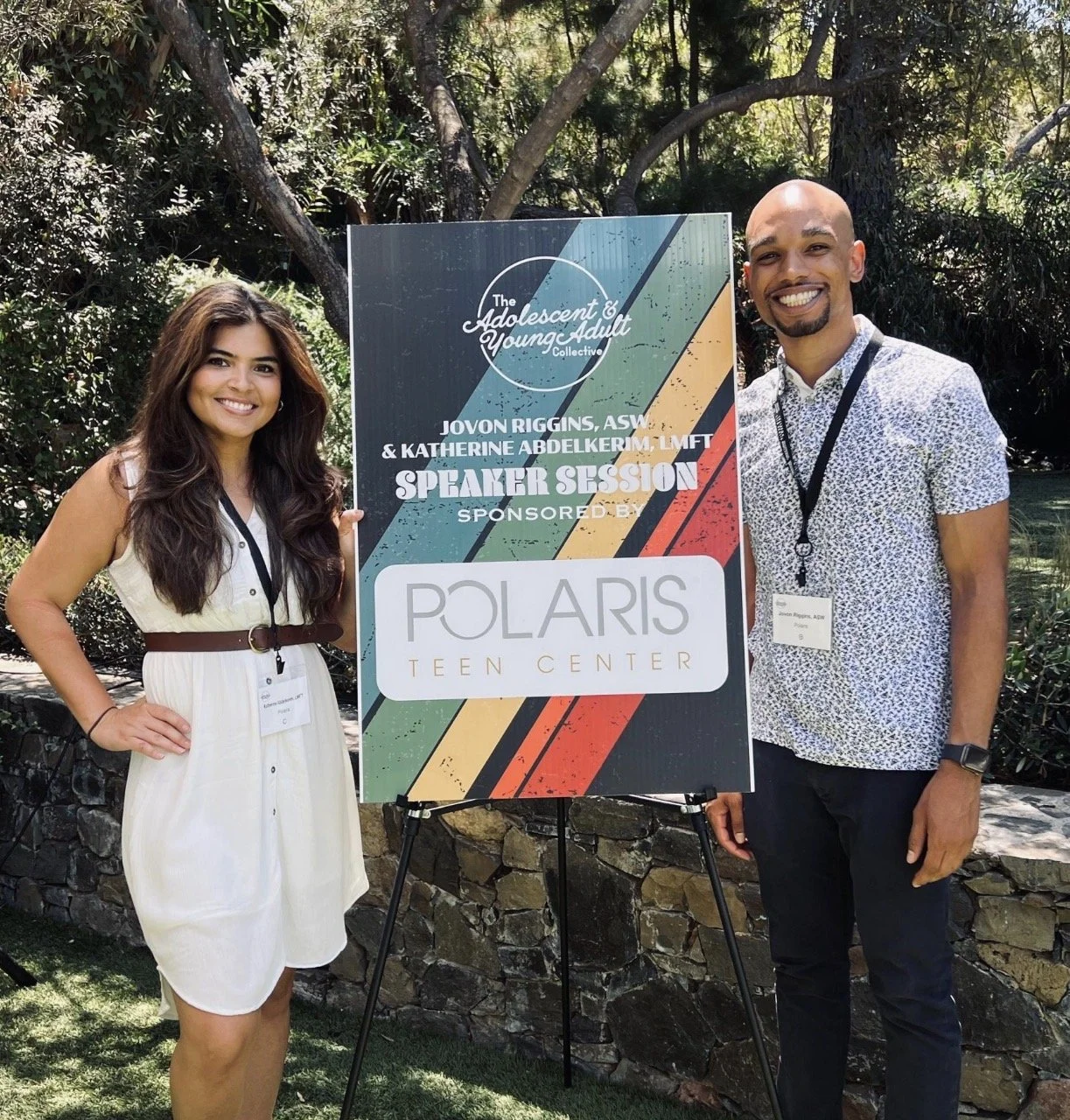Two people standing next to a colorful conference sign outdoors. The woman on the left has long brown hair and is wearing a white dress with a brown belt. The man on the right has a shaved head, goatee, and is wearing a short-sleeve shirt with a lany