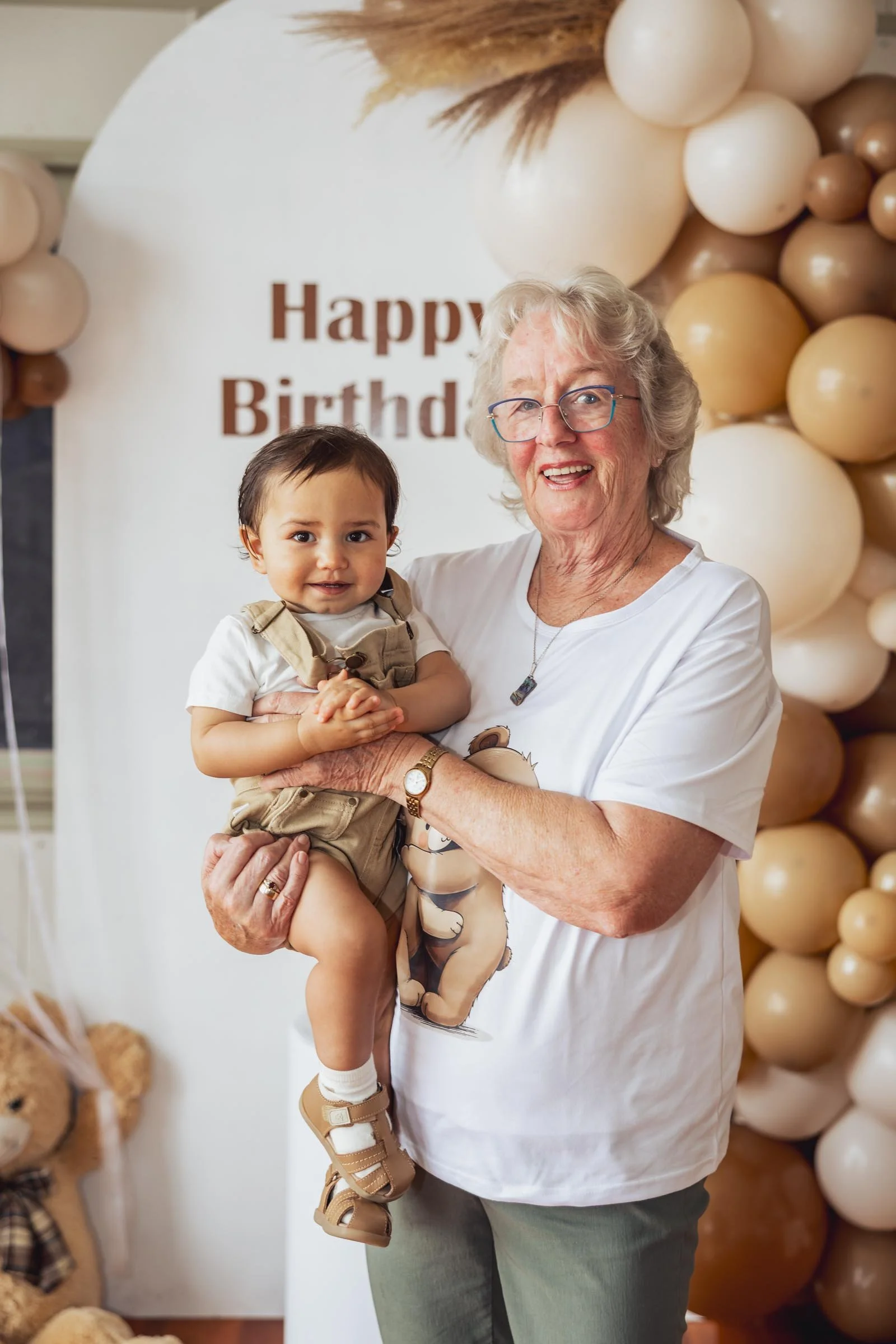 A woman with gray curly hair and glasses holding a young boy with dark hair in front of a birthday backdrop with balloons and a sign that says 'Happy Birthday'.