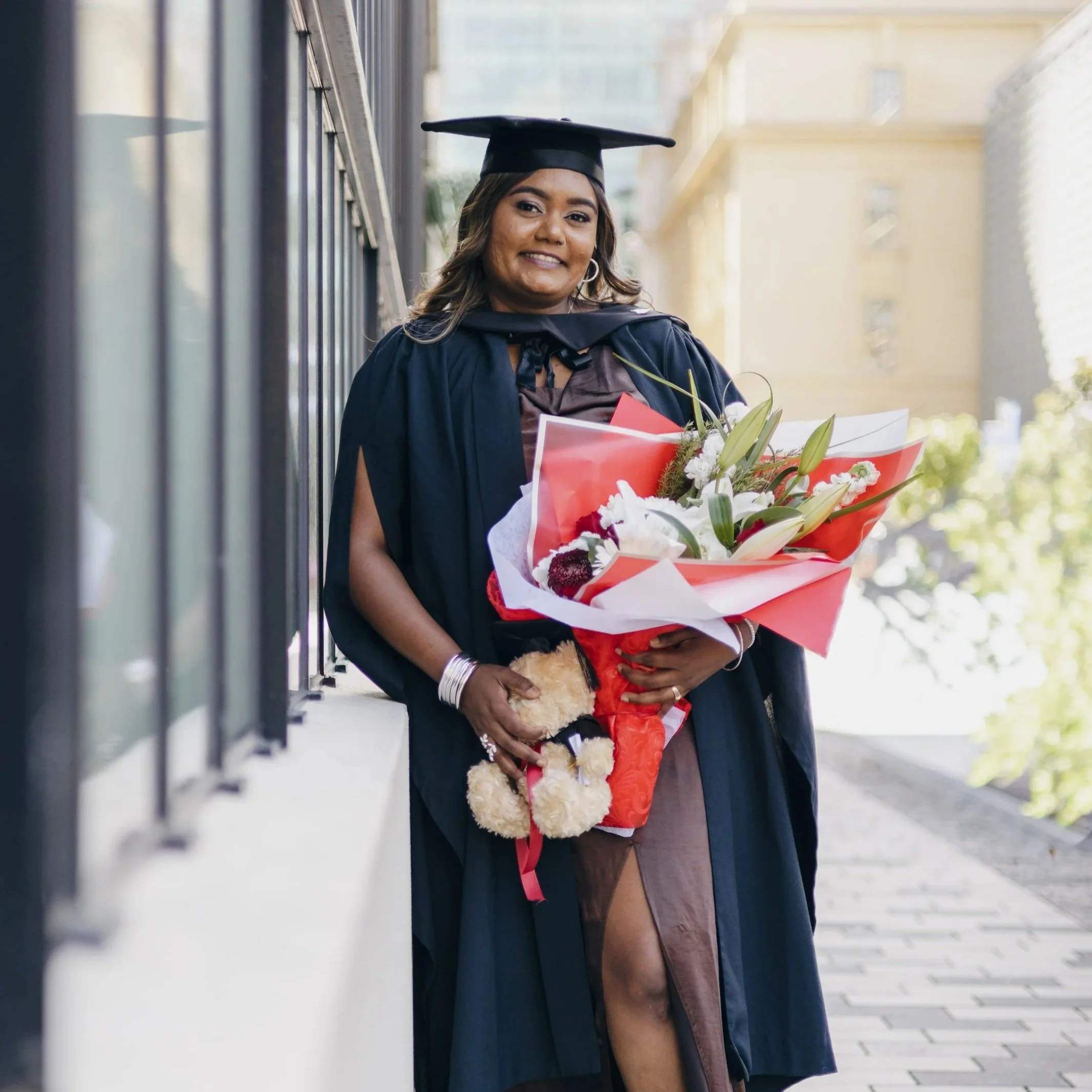 A woman wearing a graduation cap and gown holding a bouquet of flowers, standing outdoors near a building. She is smiling at the camera.