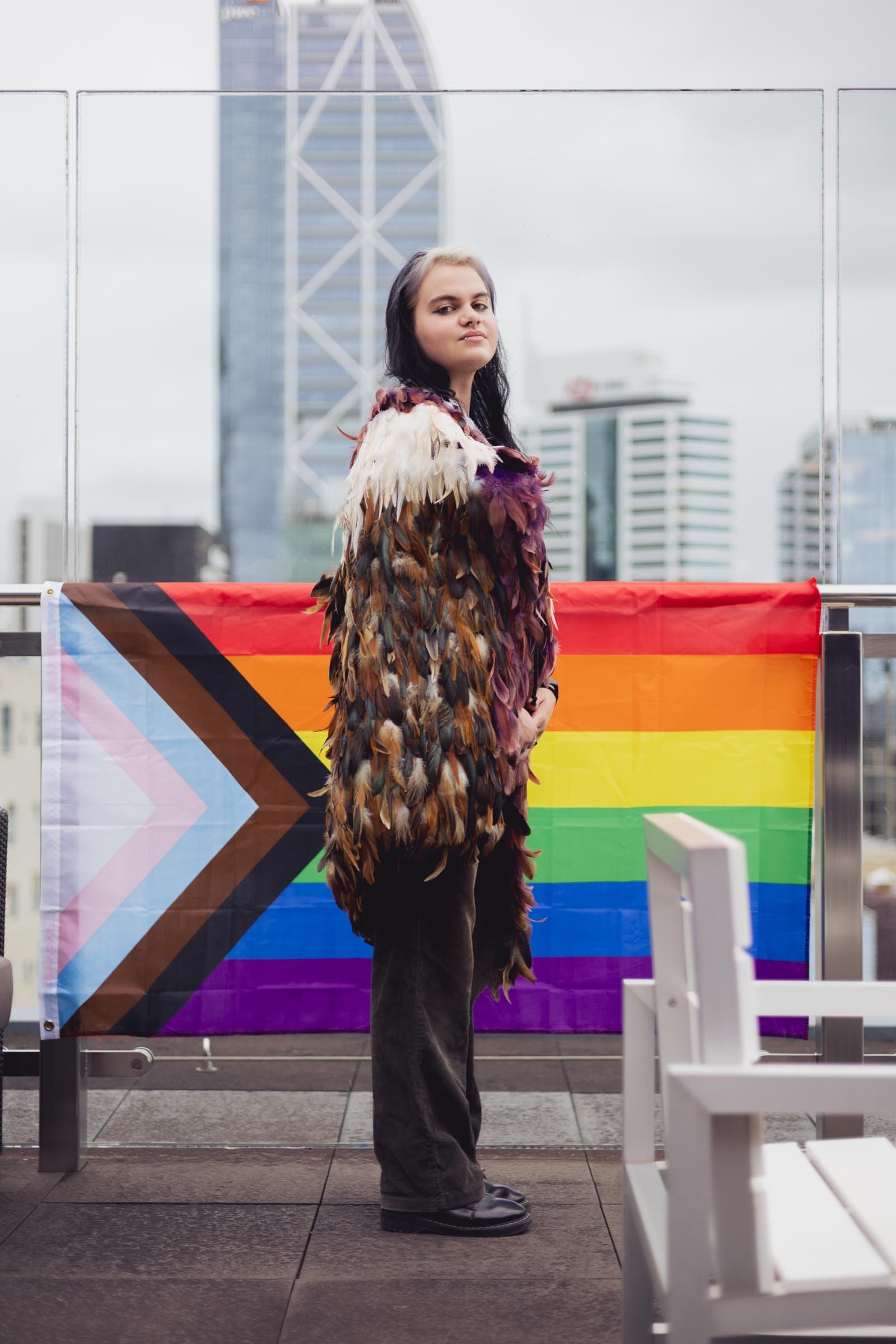 Person with long dark hair wearing a feathered multicolored coat and black pants standing on a rooftop with a city skyline in the background, including a rainbow pride flag hanging behind them.