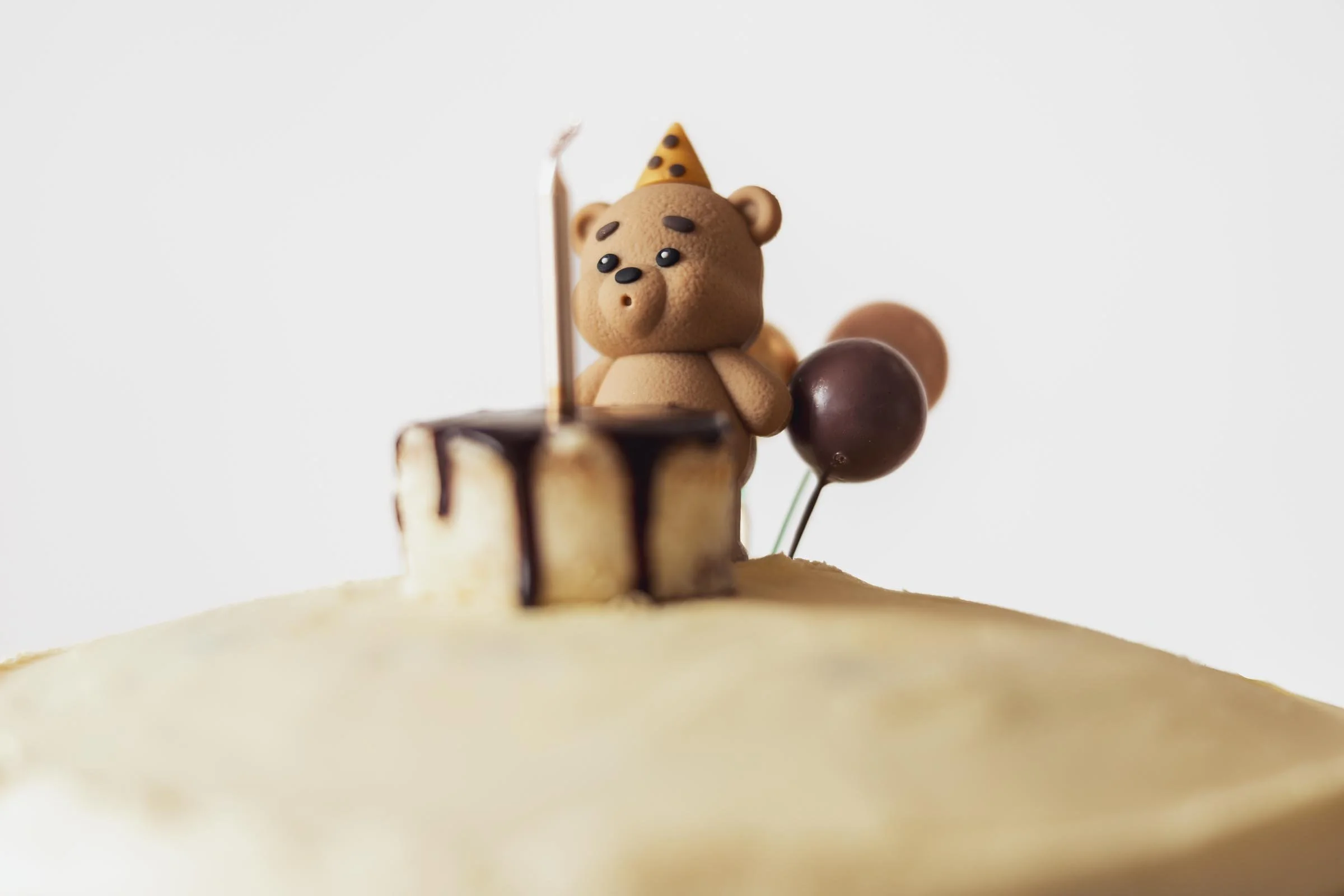 A cake decorated with a bear figurine wearing a party hat and chocolate drizzles, with balloons in the background.