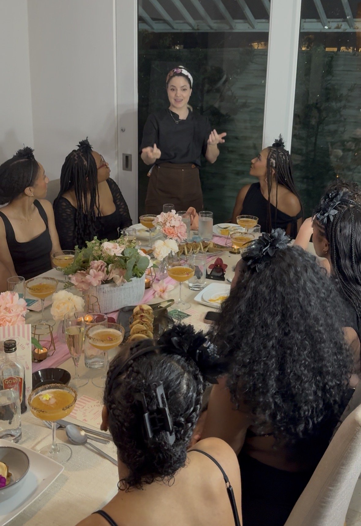 A woman standing and speaking to a group of women seated around a dining table, with floral decorations and food and drinks on the table, indoors with large glass windows behind.