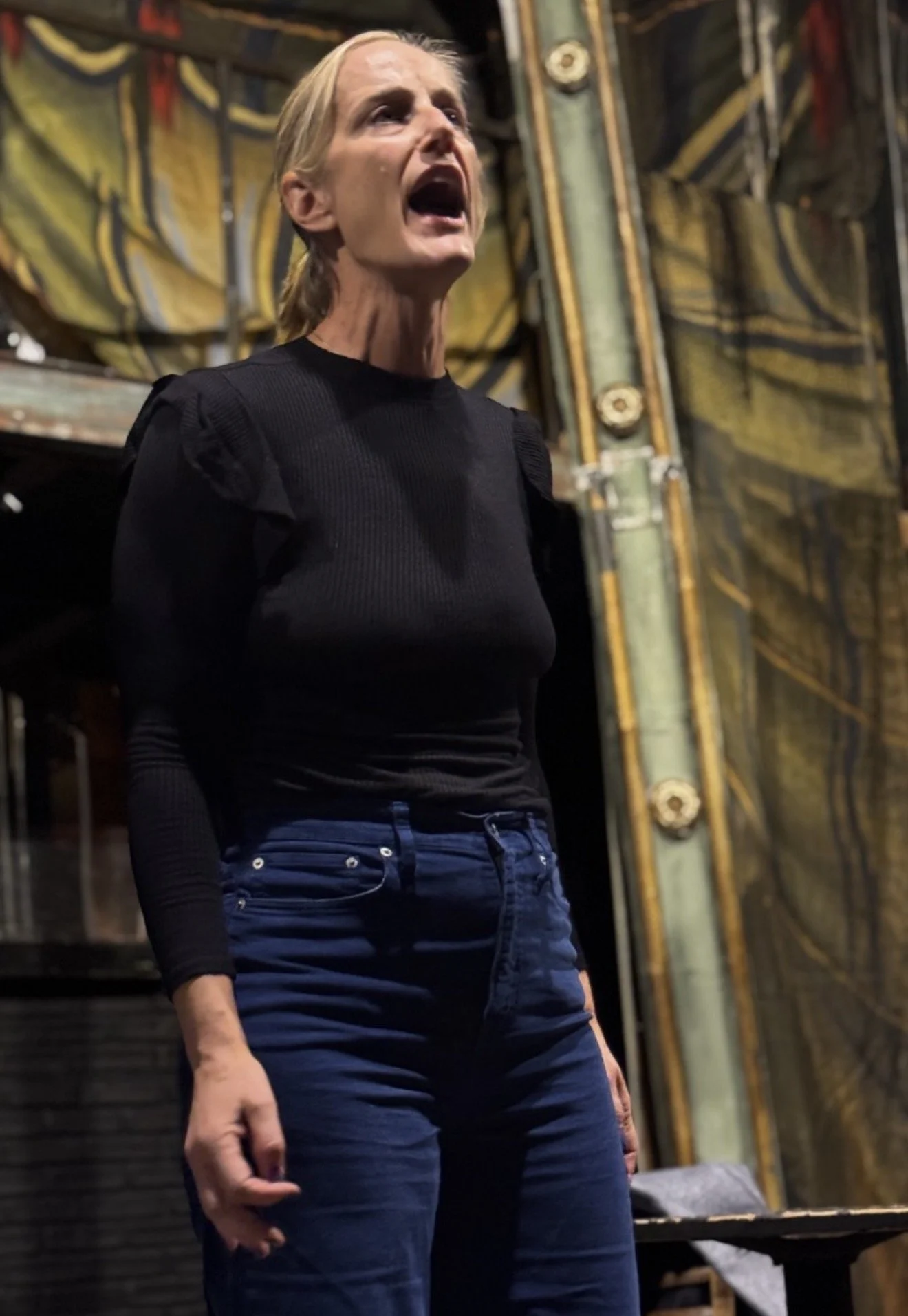 A woman with blonde hair singing or speaking passionately on a stage with elaborate gold and dark background decorations.