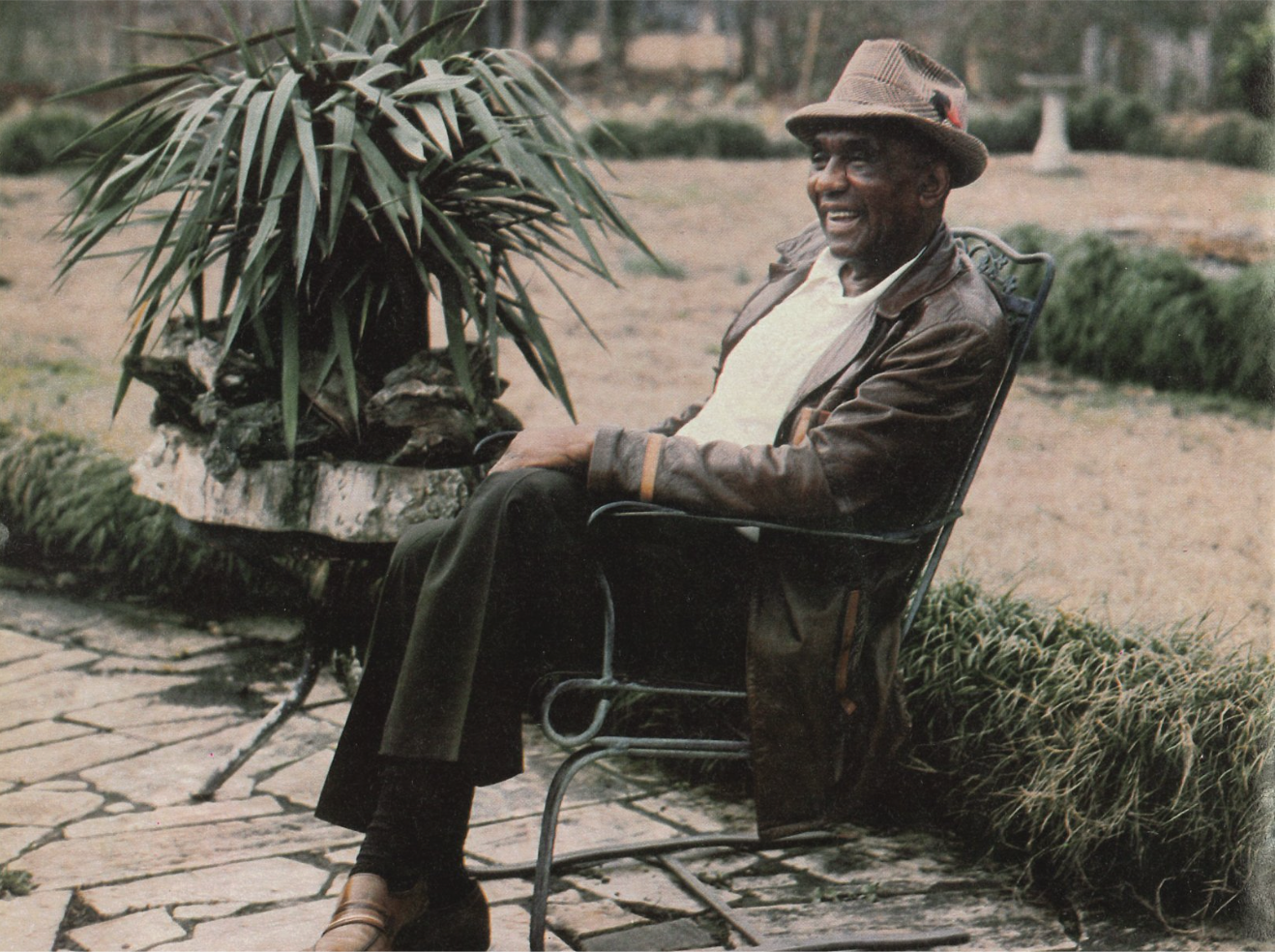 An elderly man wearing a brown hat, brown leather jacket, and white shirt sitting on a black metal bench outside in a garden area, smiling.