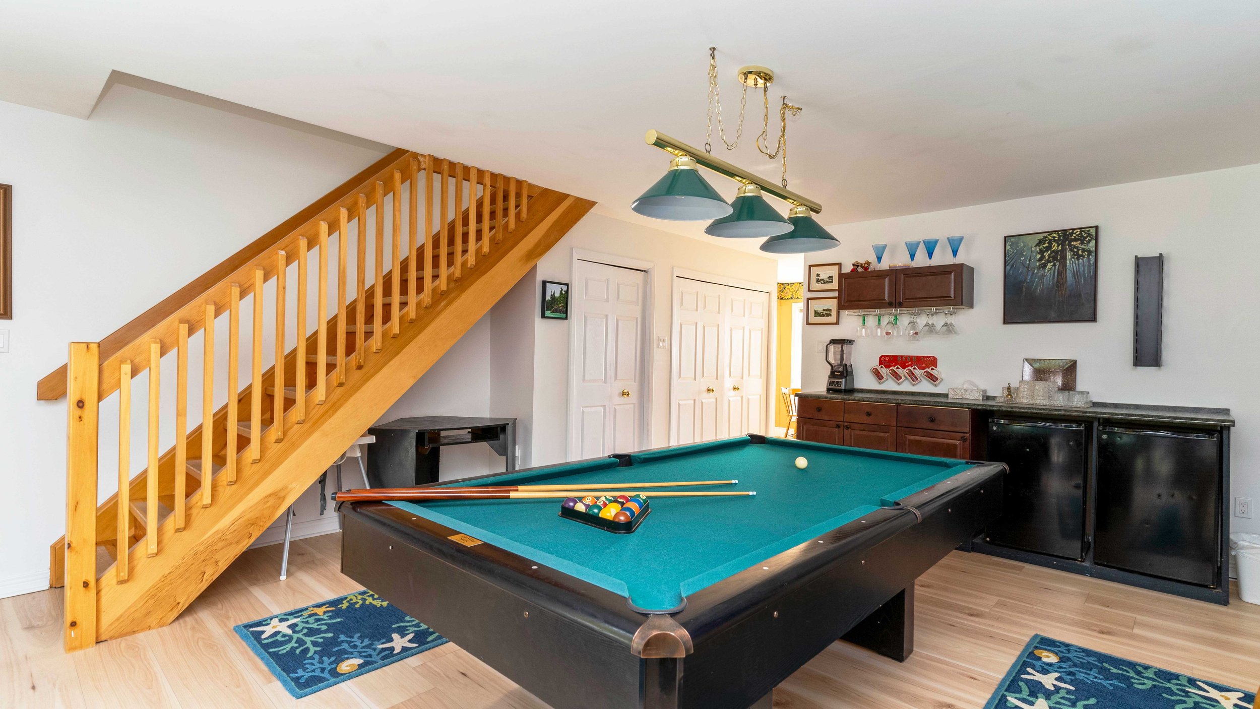 Basement game room with a pool table, cues, and billiard balls. Wooden staircase, bar area with glasses and blender, and decorative items on shelves. Ceiling lights and art on the walls.
