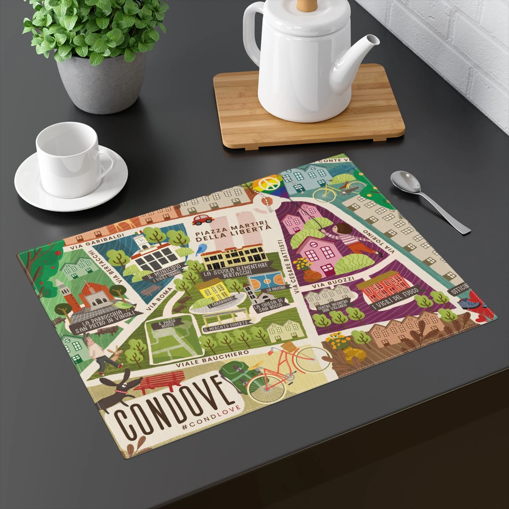 Tavola con una mappa illustrata di Condove, un bicchiere di porcellana bianca con piattino, una teiera bianca su un tagliere di legno, un cucchiaino di metallo, una pianta verde in un vaso di cemento, sul tavolo scuro con parete bianca sullo sfondo.