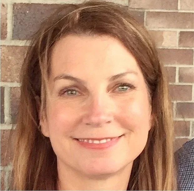 Close-up of a woman with brown hair and a friendly smile standing in front of a brick wall.