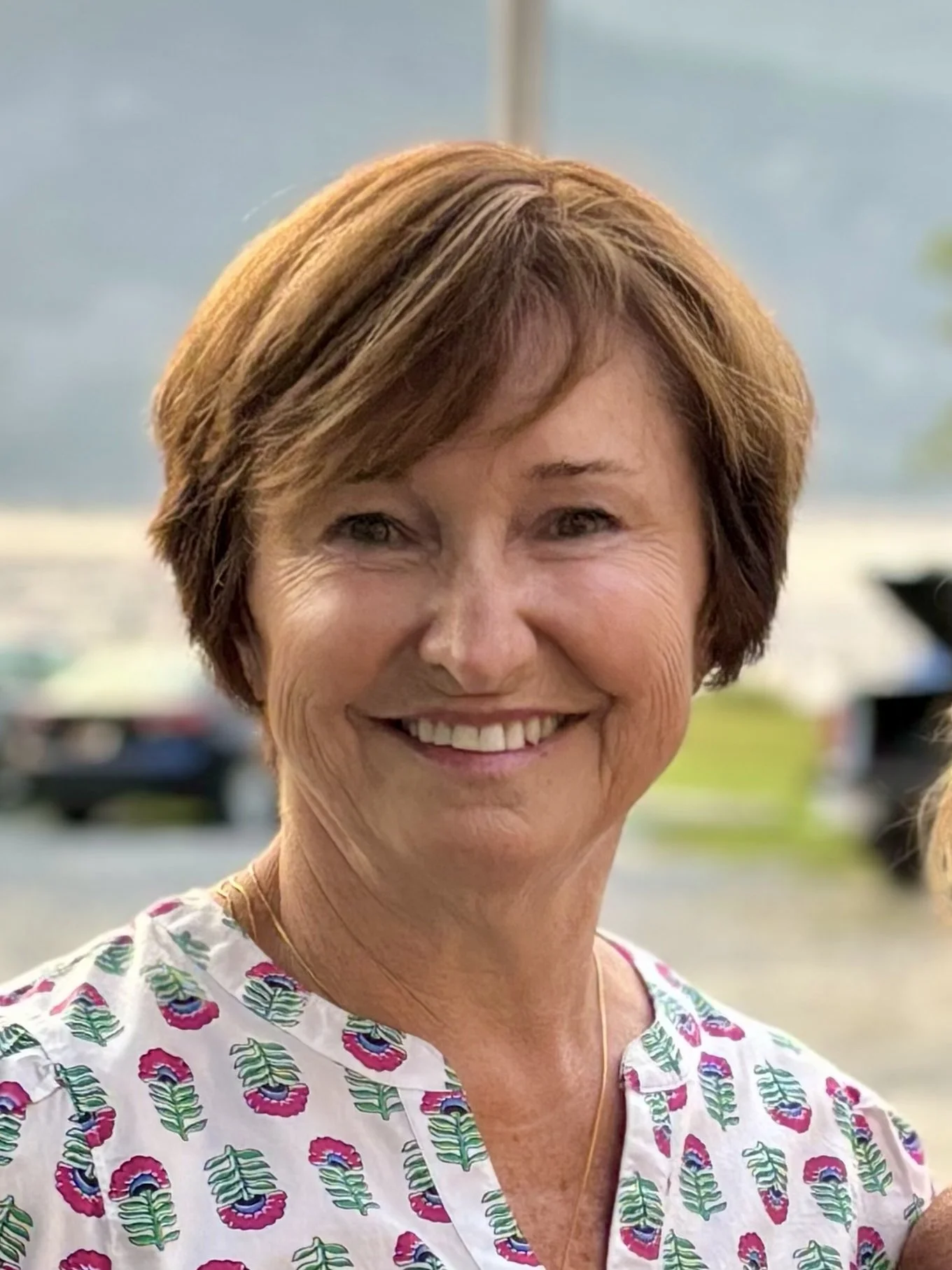 A smiling woman with short brown hair wearing a white shirt with colorful floral patterns, outdoors with a blurred background of cars and trees.